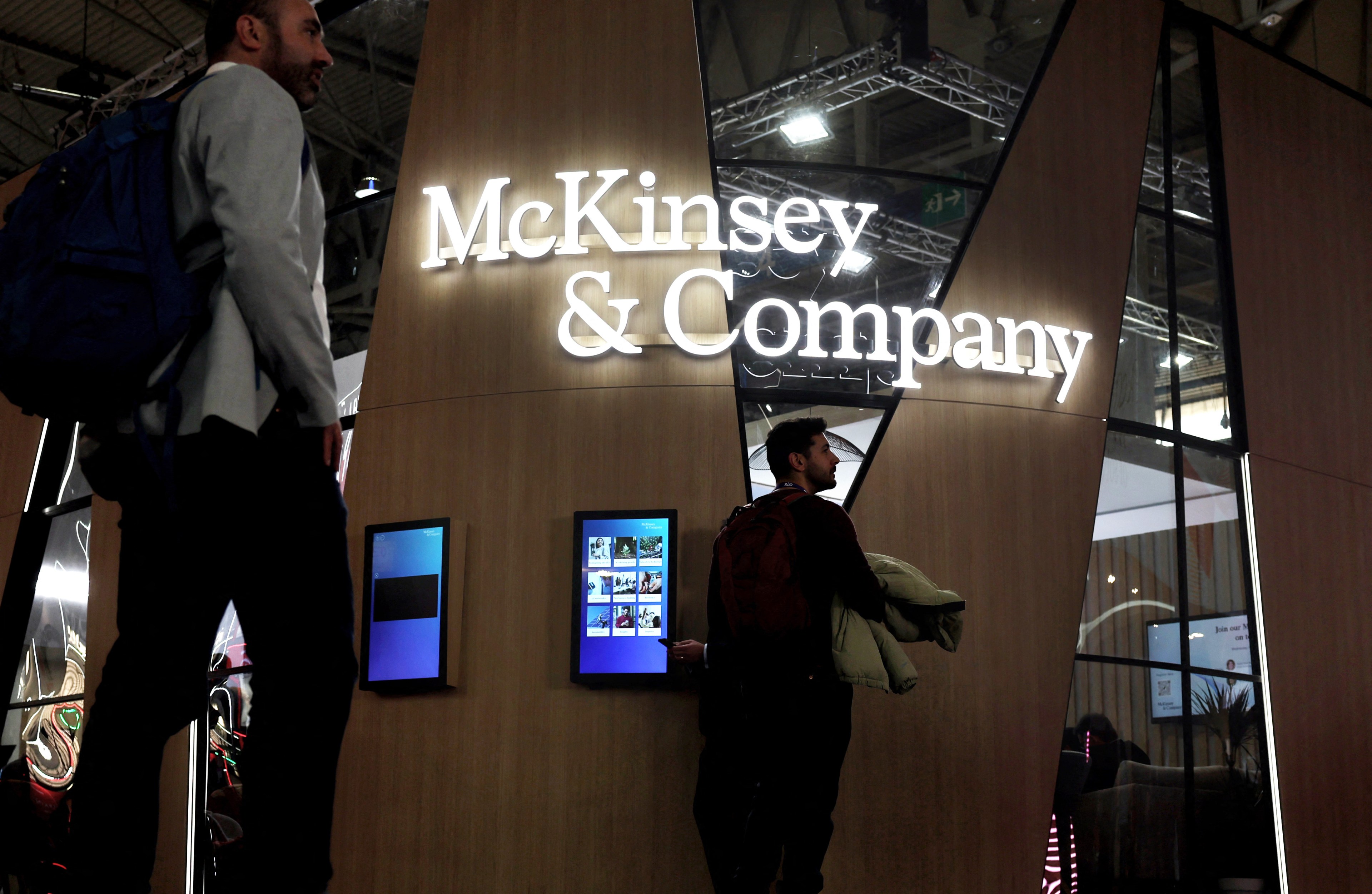 A booth with &quot;McKinsey &amp; Company&quot; in bright letters on a wooden panel. Two men stand nearby, one looking at a touchscreen display.