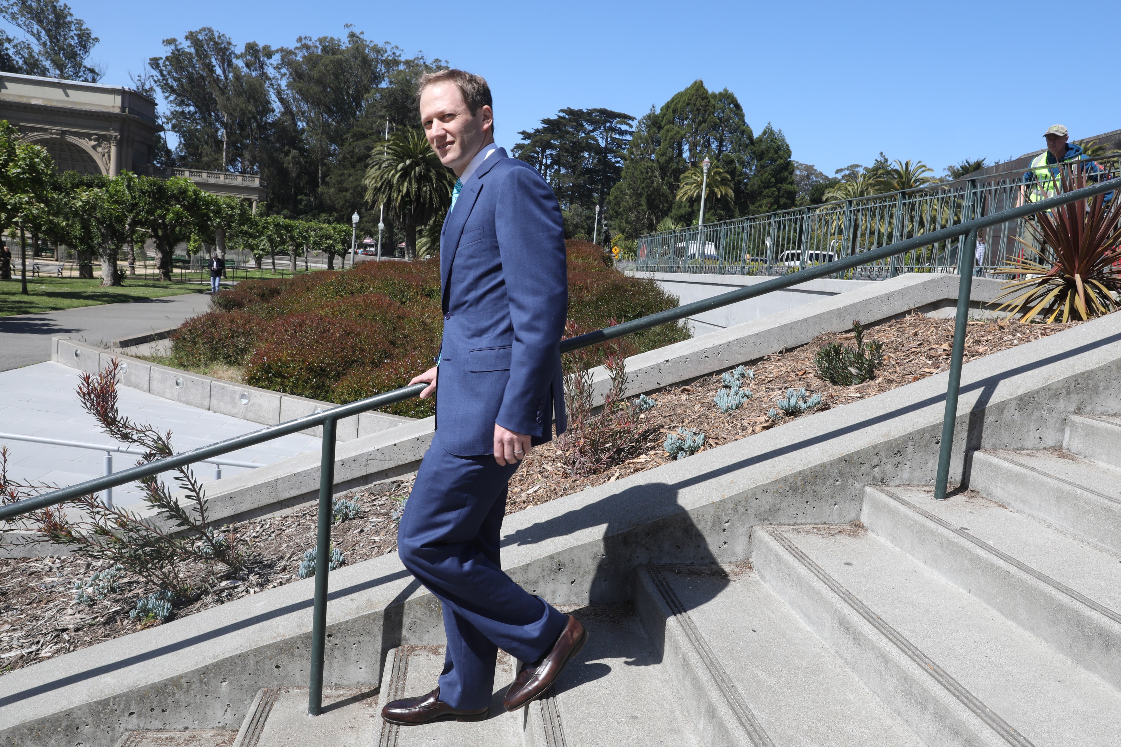 A man in a blue suit walks up concrete steps with greenery and trees around. The sky is clear and blue, and a railing follows the stairs.