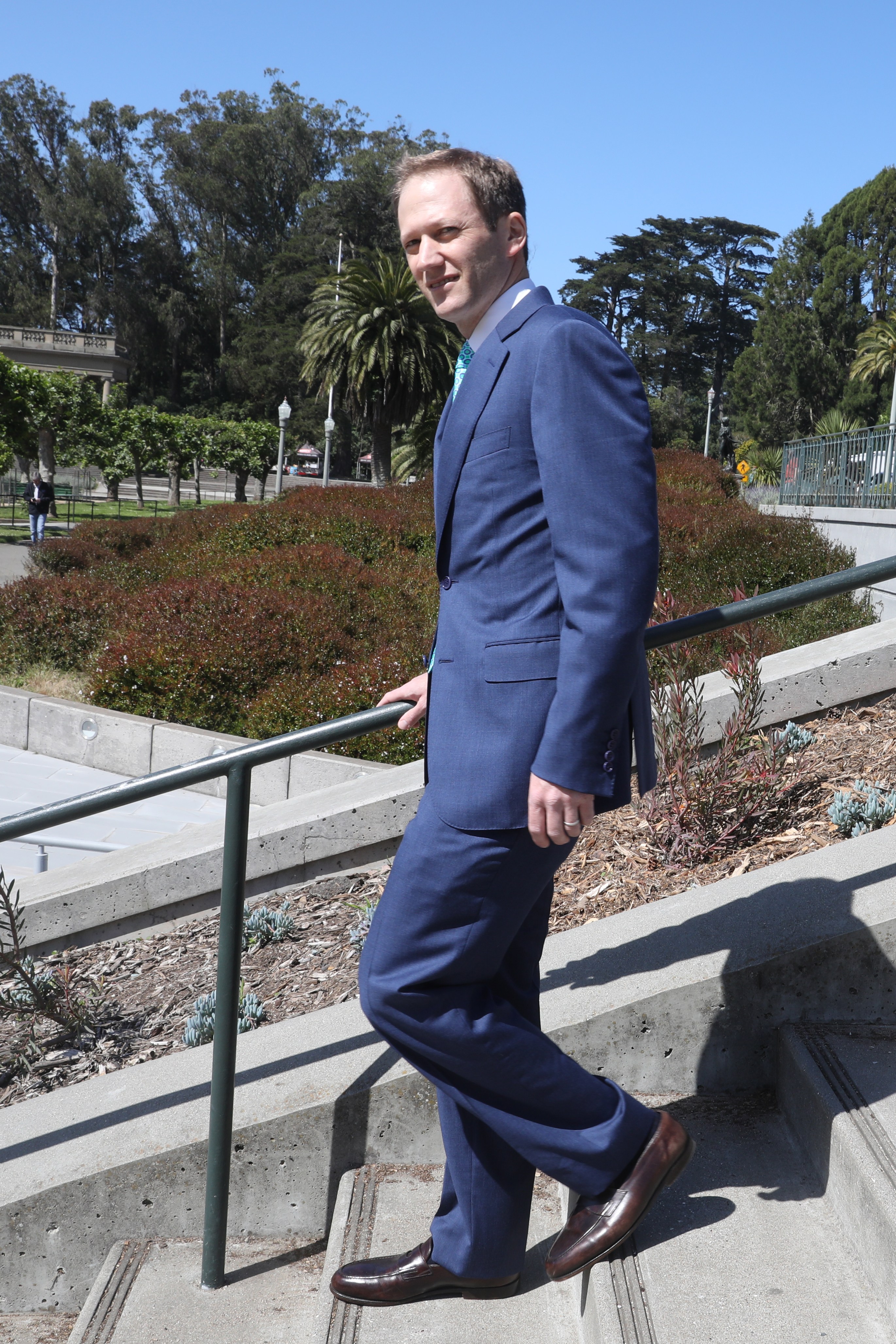 A man in a blue suit walks up concrete steps with greenery and trees around. The sky is clear and blue, and a railing follows the stairs.