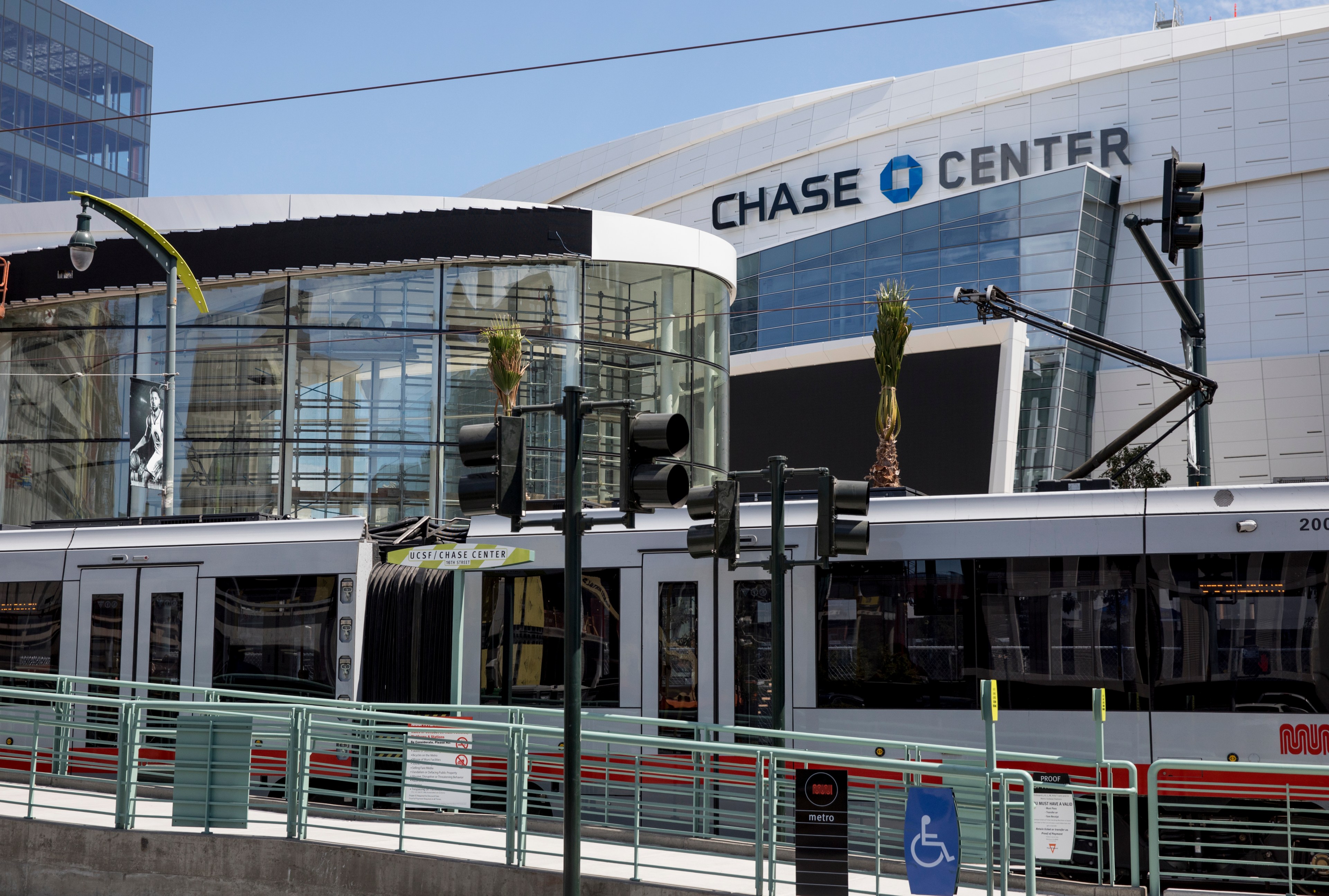 A modern tram passes in front of the Chase Center, which features a sleek glass and steel design. A UCSF/Chase Center sign is visible, and palm trees are planted nearby.