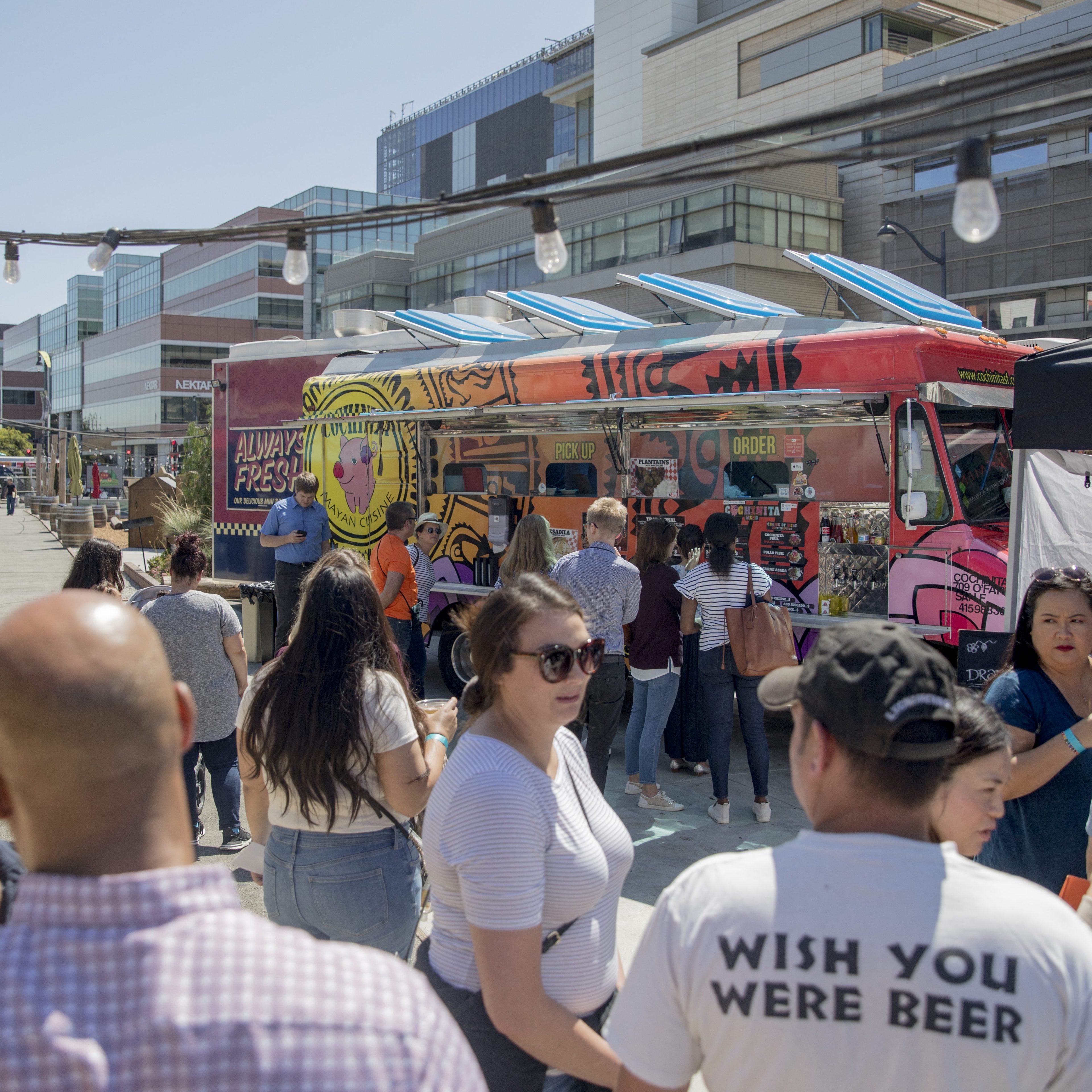 A colorful food truck with a long line of people is stationed in a bustling urban area, surrounded by modern buildings under a sunny sky.