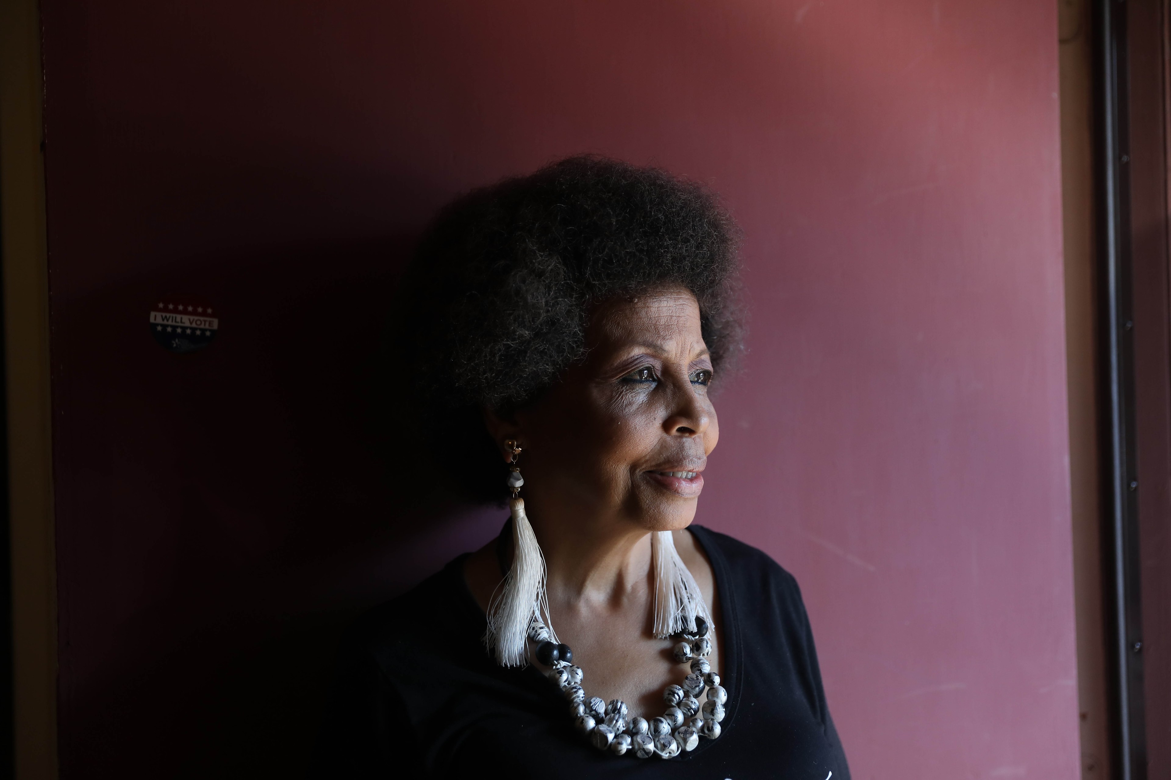 The image shows a thoughtful woman with a natural hairstyle looking to the side. She wears large earrings and a necklace, standing against a reddish-brown wall.