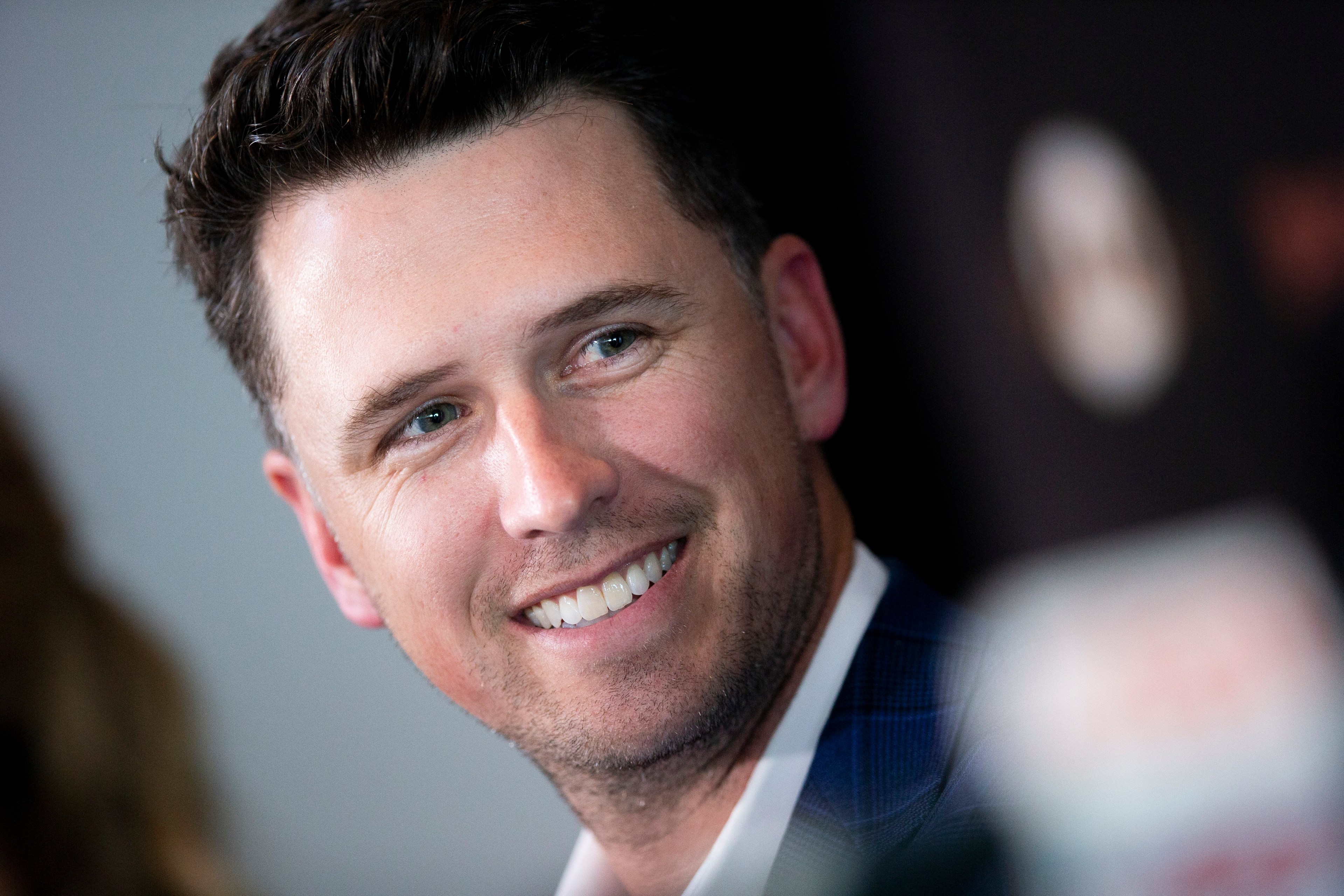 A man with short dark hair is smiling, looking to the side. He is wearing a suit, and the background is blurred with some indistinct objects.