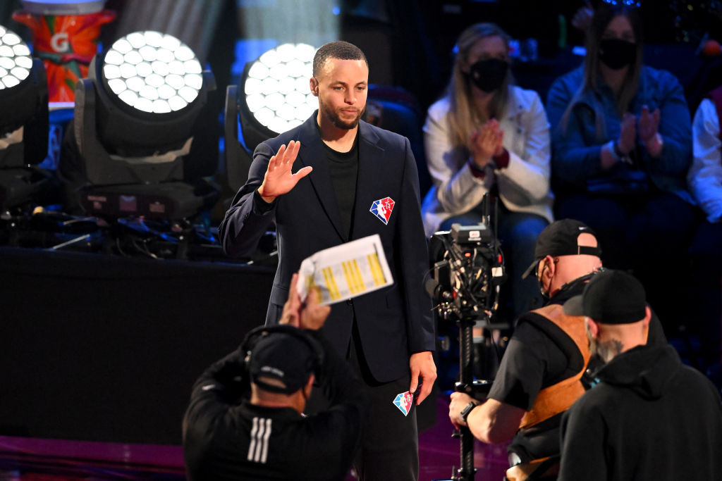 A man in a dark blazer waves to the audience, standing amid bright stage lights. Behind him, spectators applaud. Camera crew capture the moment in front.