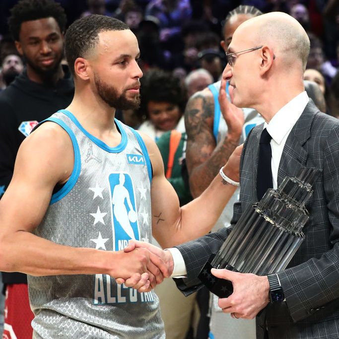 A basketball player in a gray jersey shakes hands with a man in a checkered suit holding a trophy. Others are clapping in the blurred background.