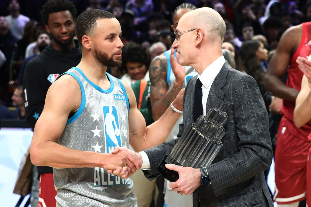 A basketball player in a gray jersey shakes hands with a man in a checkered suit holding a trophy. Others are clapping in the blurred background.