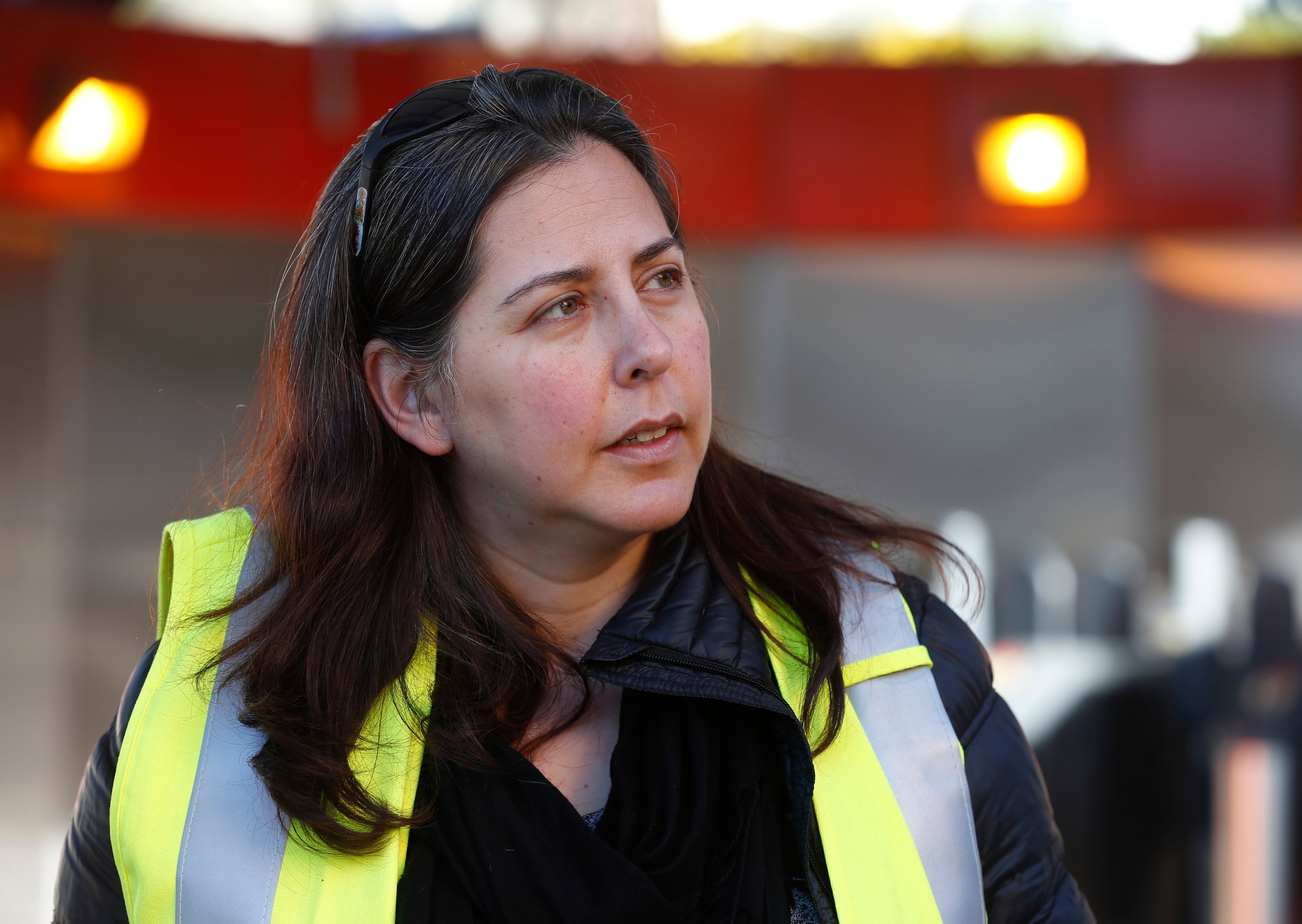 A woman with long, dark hair wears a reflective safety vest and a black jacket. She looks to the side, with a blurred background featuring bright lights.