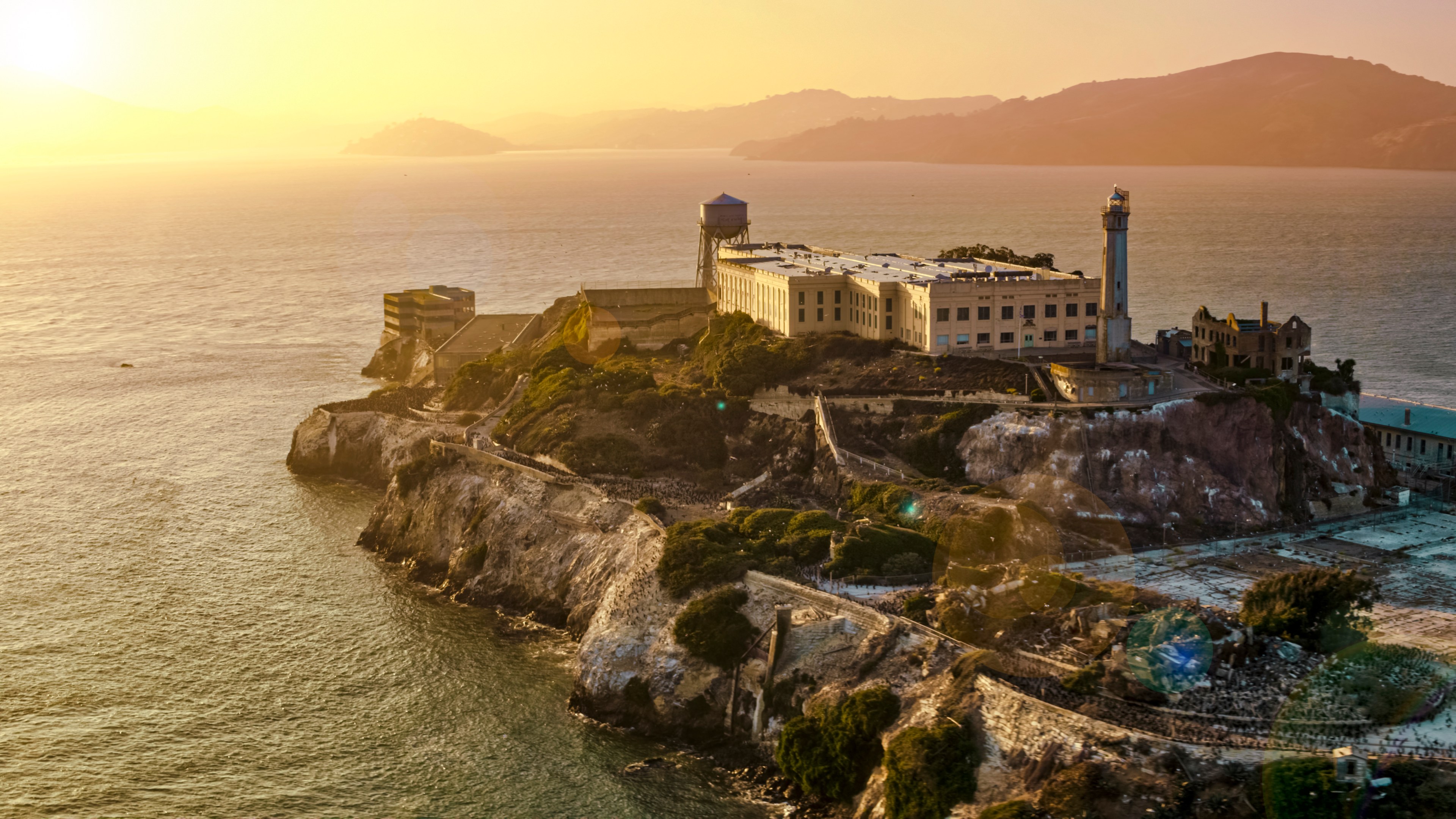 The image shows Alcatraz Island at sunset, with its famous prison building and lighthouse. The island is surrounded by water and a golden sky.