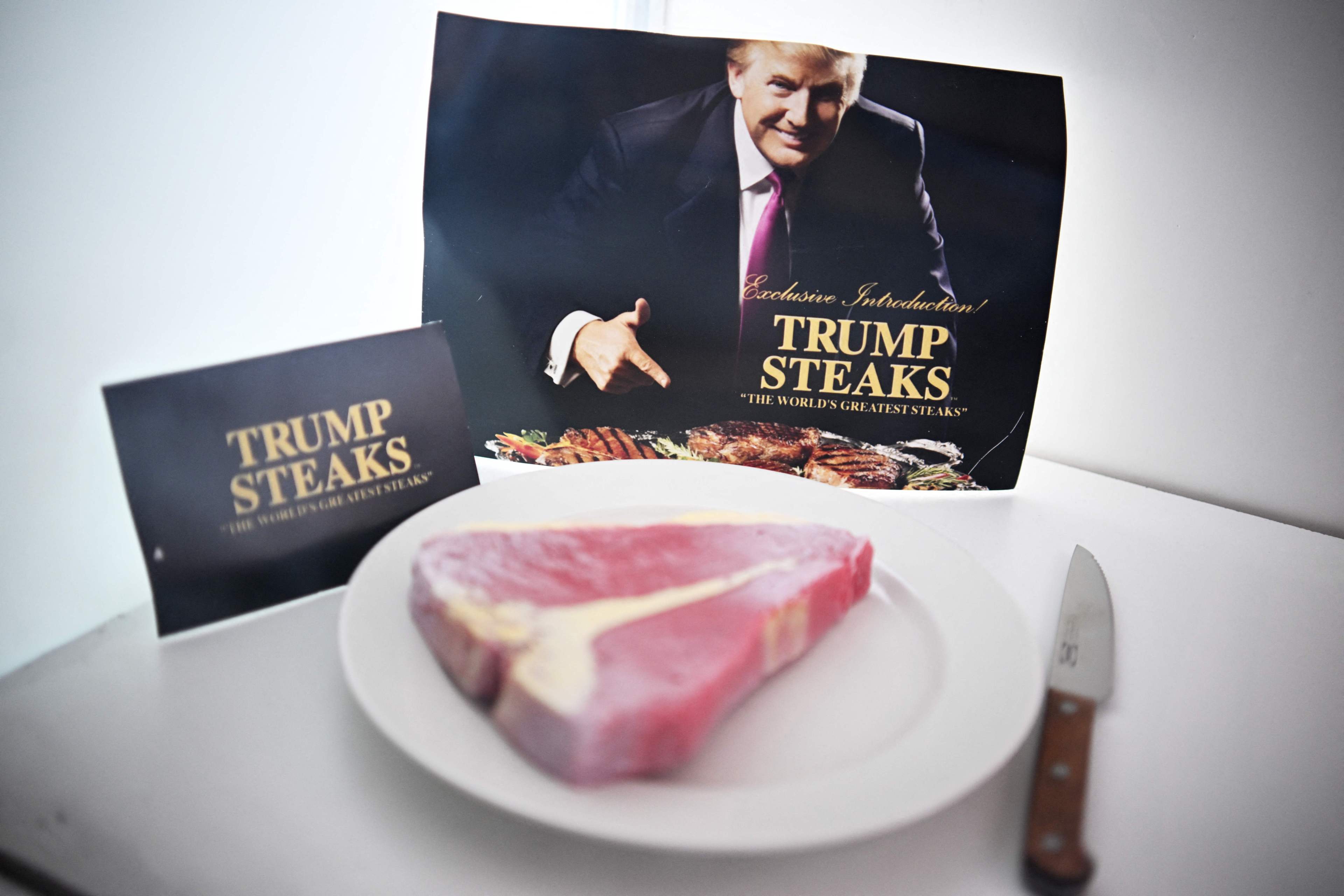 A raw steak sits on a white plate beside a knife. Behind it are two signs promoting "Trump Steaks" as "The World's Greatest Steaks."