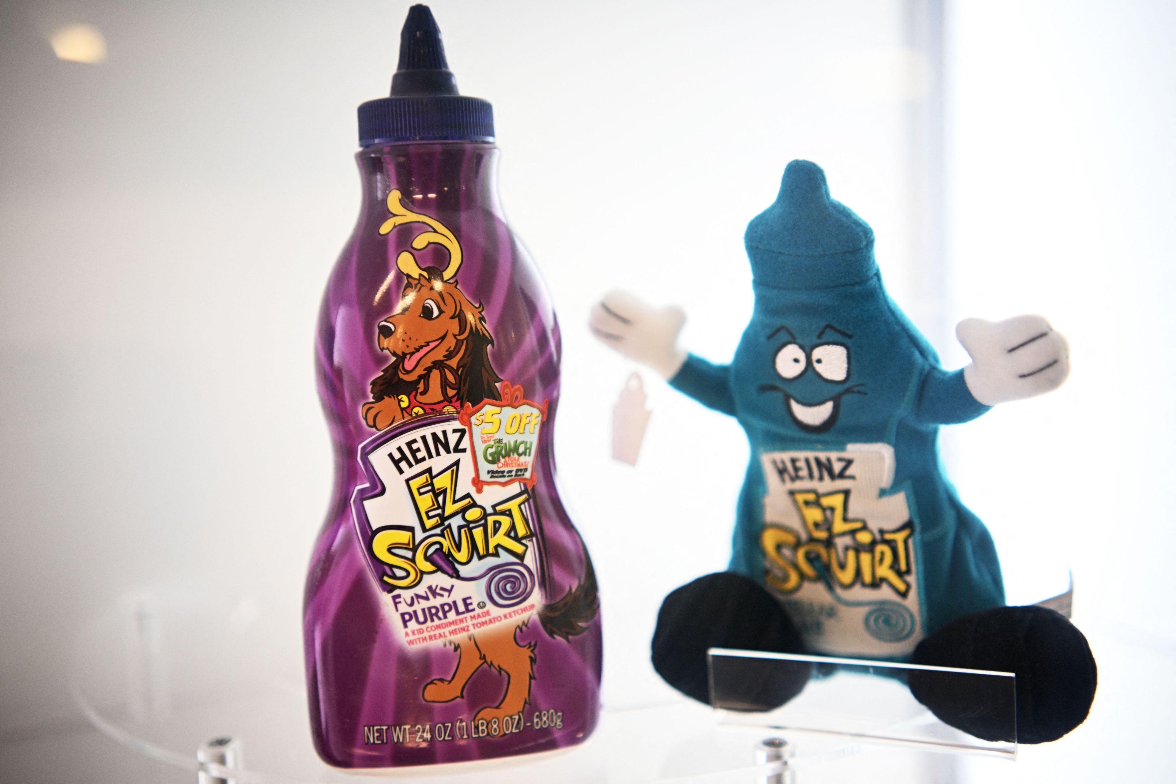 A purple Heinz EZ Squirt ketchup bottle labeled "Funky Purple" next to a blue plush toy shaped like a ketchup bottle with a cheerful face.