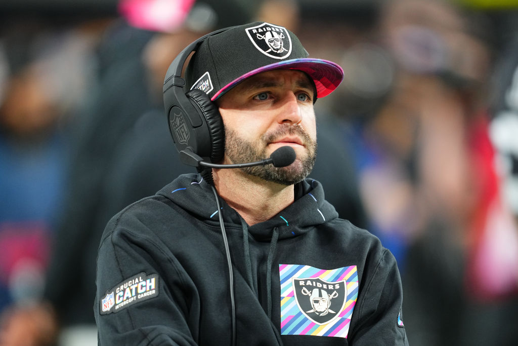 A man wearing a Raiders cap and a headset is focused, possibly coaching during a game. He wears a black hoodie with a colorful Raiders logo.