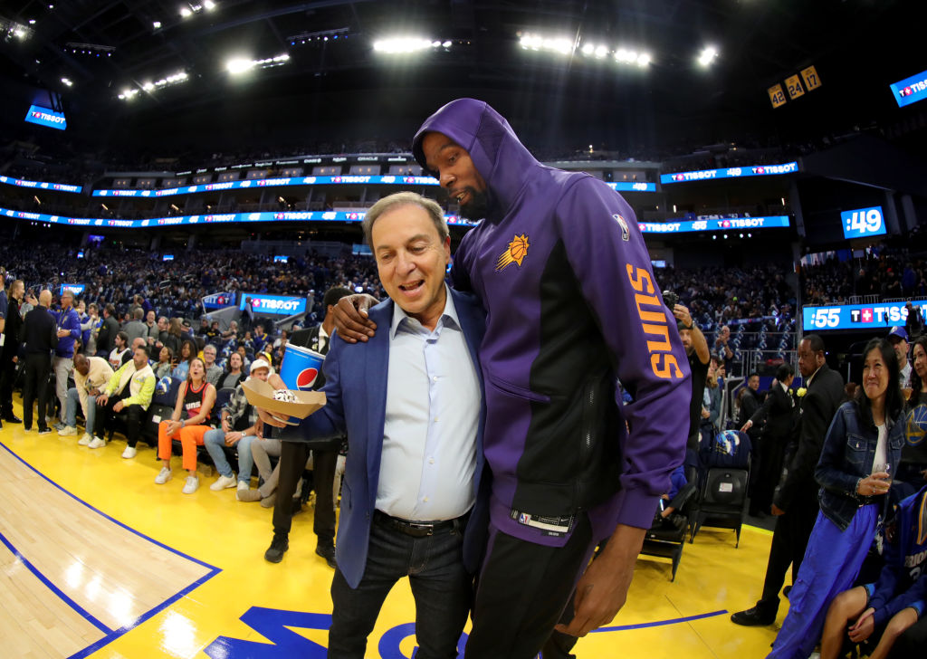 A man in a purple &quot;Suns&quot; jacket has his arm around another man holding a drink and food, standing courtside at a basketball game with a crowd behind them.