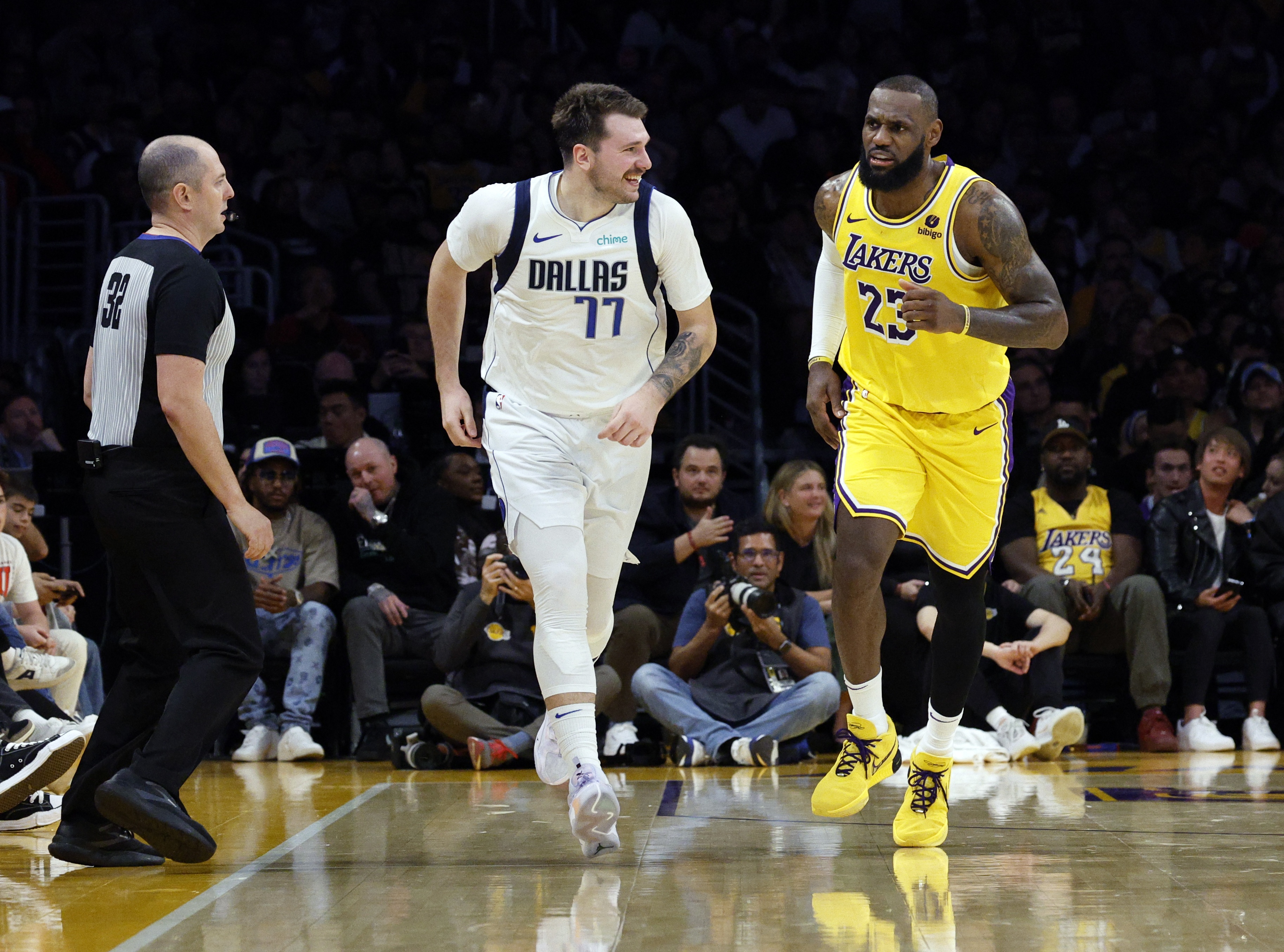 Two basketball players, one in a Dallas Mavericks jersey and the other in a Los Angeles Lakers jersey, are on the court with a referee walking beside them.