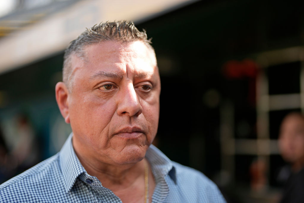 A man with short hair and a serious expression stands outdoors. He wears a checkered shirt, and the background is slightly blurred with shadowy figures and structures.