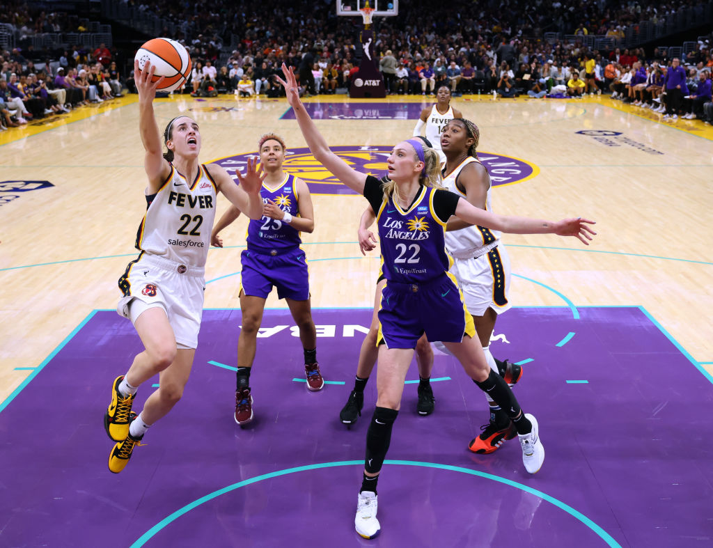 A basketball player in a white Fever uniform jumps to attempt a shot, while three players in purple Los Angeles uniforms defend, on a crowded court.