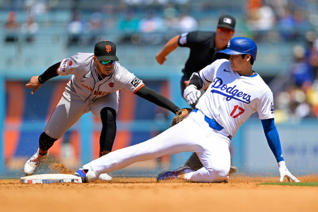 A baseball player in a Dodgers uniform is sliding into a base as a player in a Giants uniform attempts to tag him; an umpire observes closely.