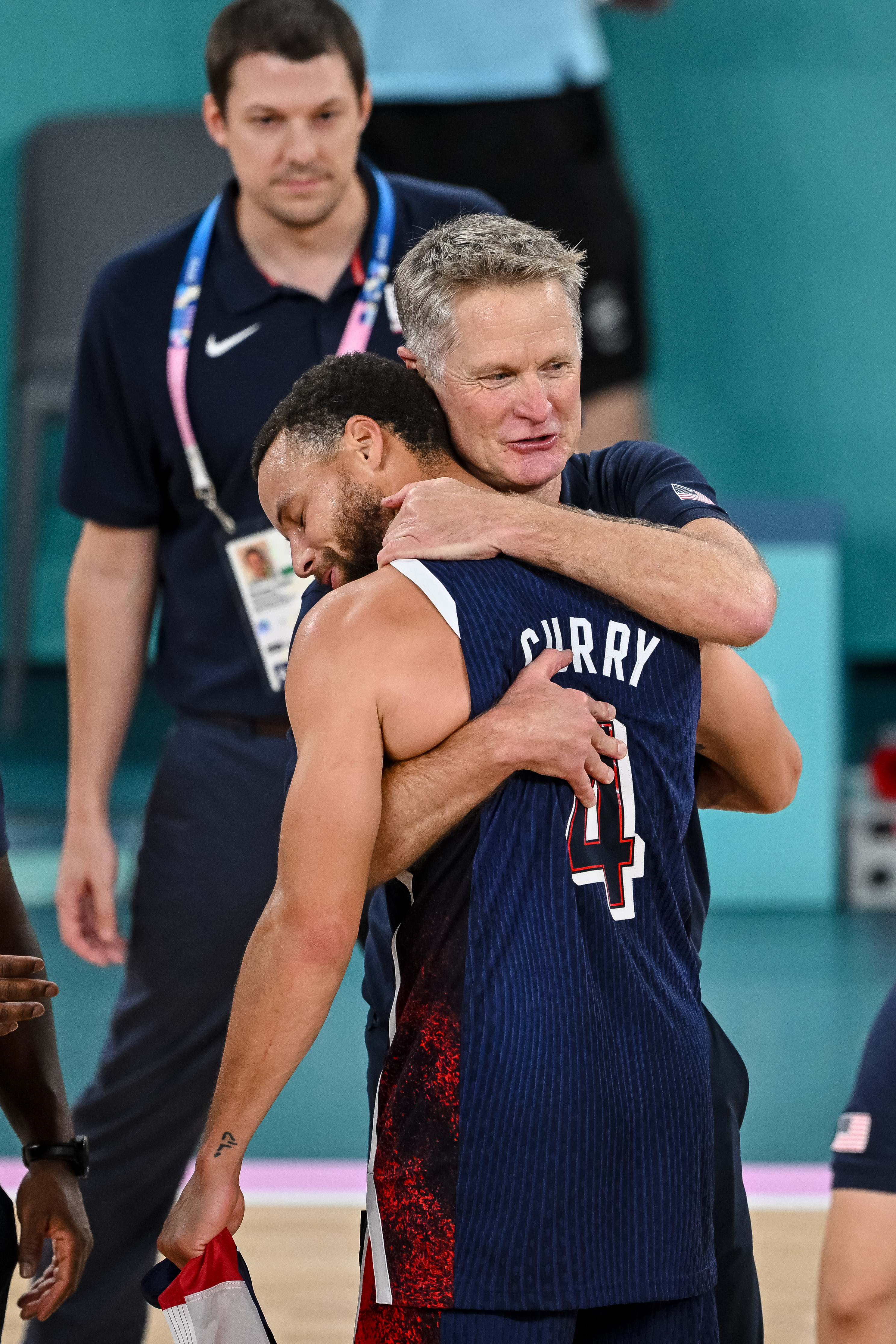 A man wearing a USA basketball uniform is being hugged by a coach. Another person with a lanyard stands in the background.