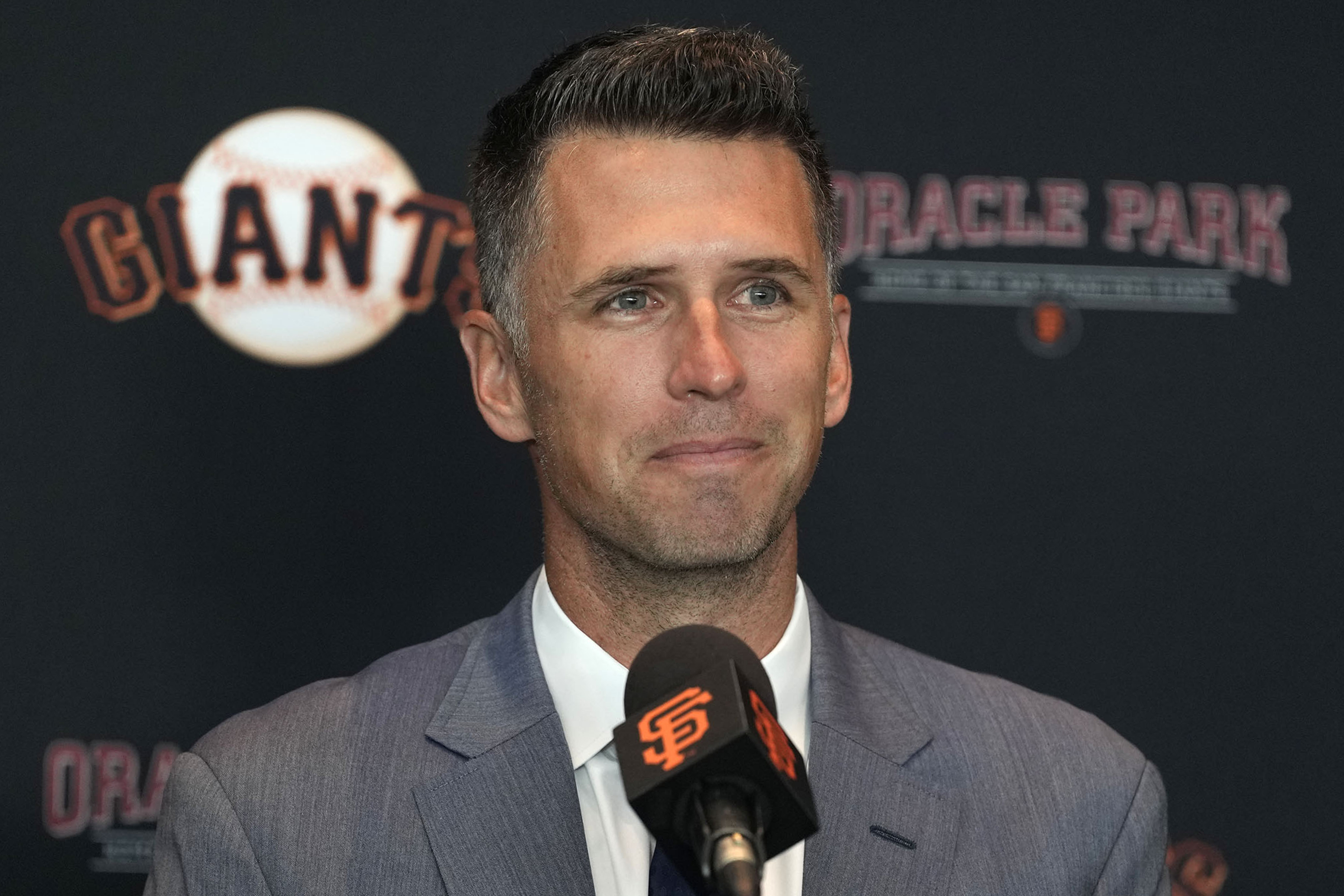 A man in a suit stands at a podium with a microphone, against a backdrop featuring "GIANTS" and "ORACLE PARK."