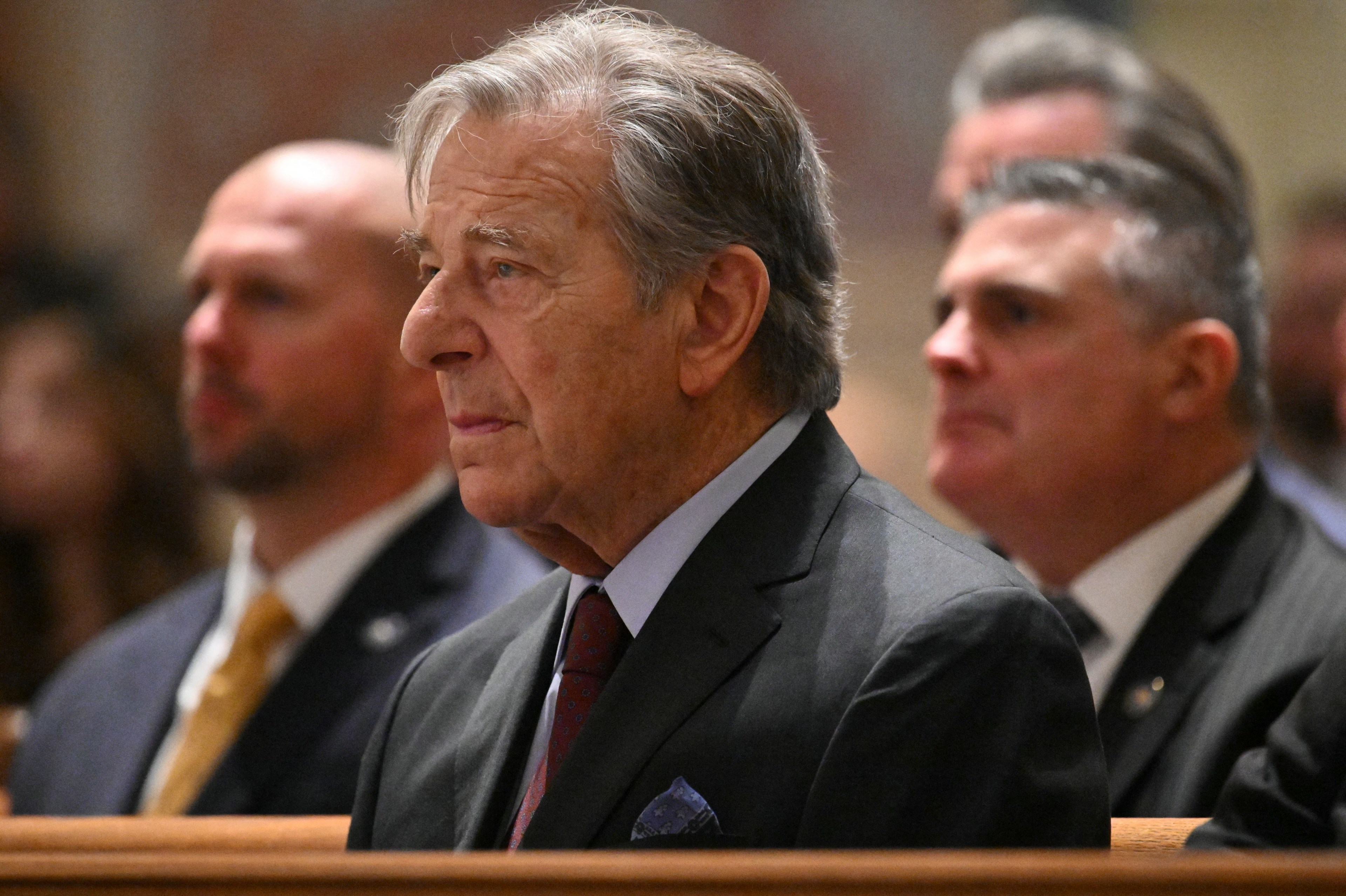 A man with gray hair is seated, wearing a dark suit and tie, looking ahead thoughtfully. Other similarly dressed individuals are visible but blurred in the background.