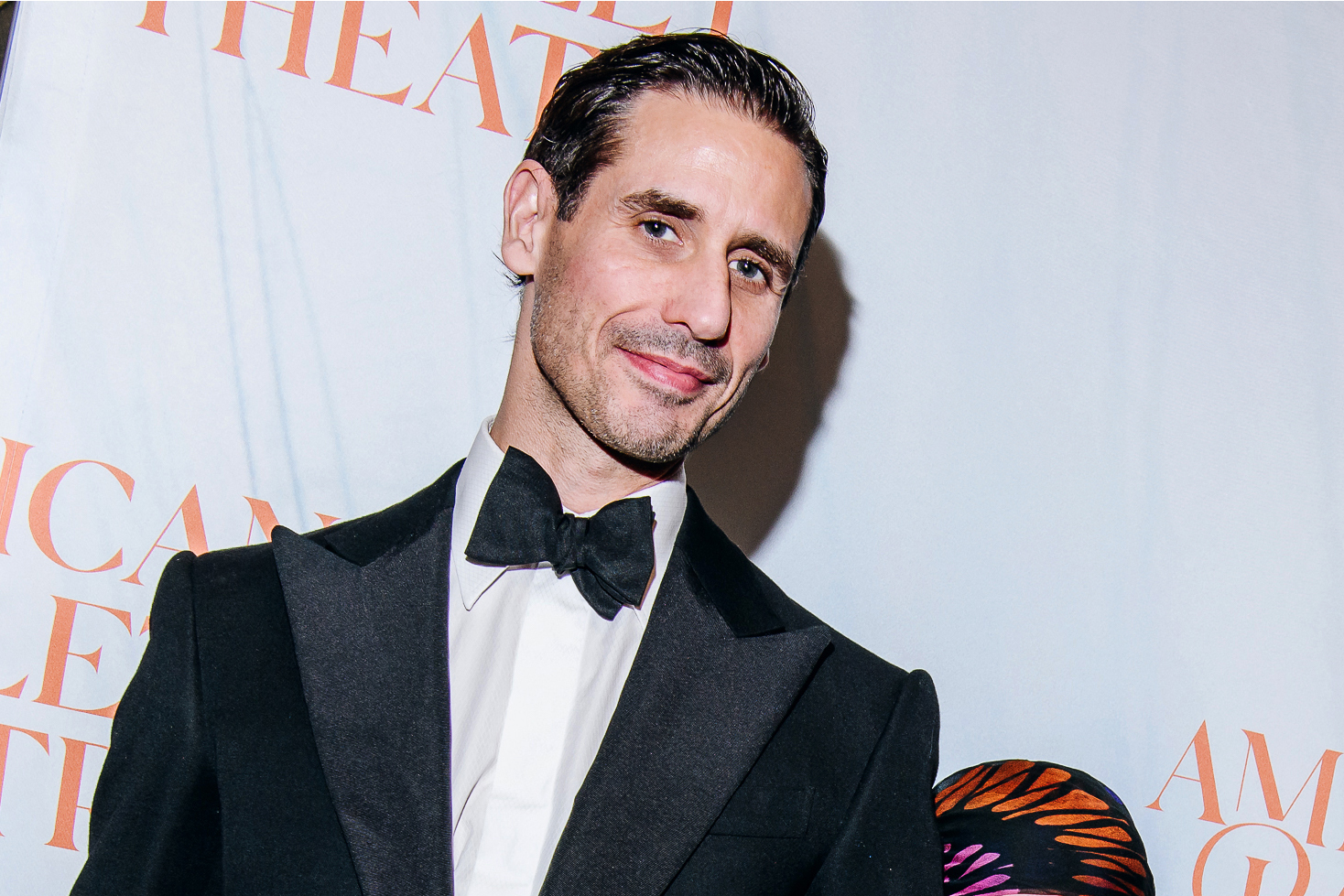 A man in a formal black tuxedo with a bow tie smiles at the camera. The backdrop features orange text partially visible, suggesting a formal event.