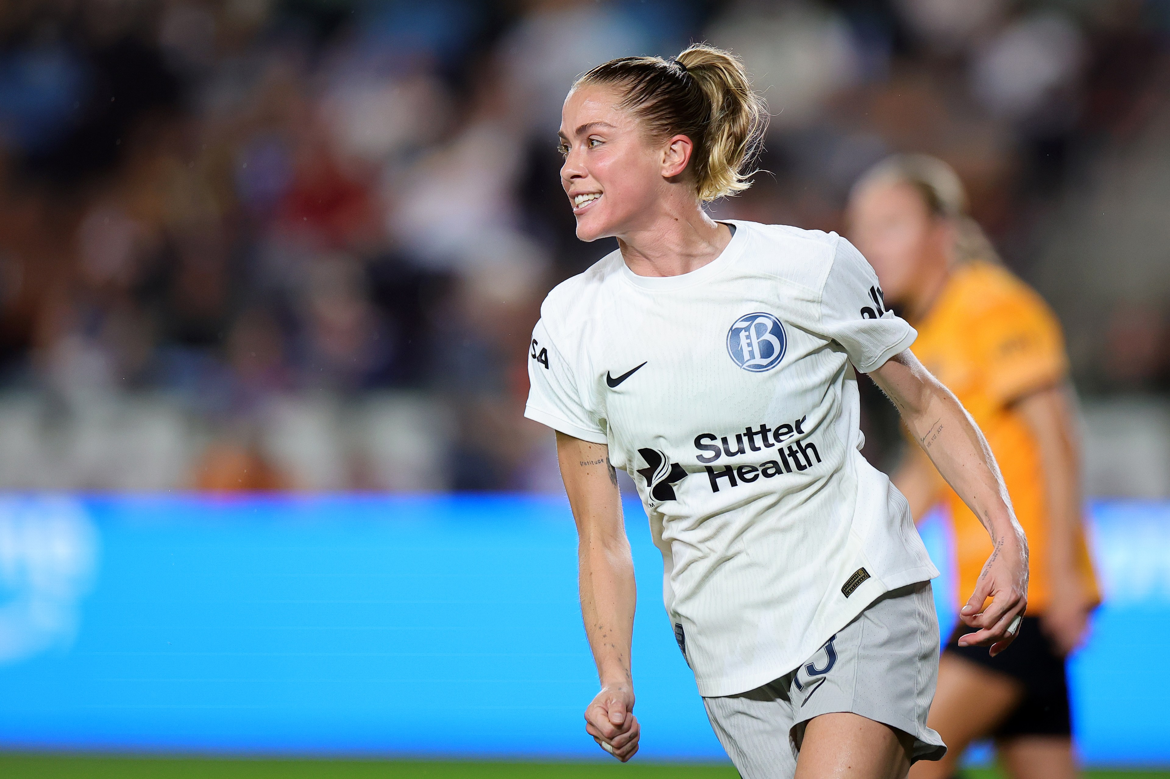 A soccer player in a white jersey runs on a field, looking energized. The jersey has blue logos, and there’s a blurred player in an orange kit behind.