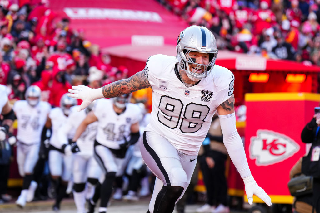 A football player in a white uniform and silver helmet enthusiastically runs onto a field, leading his team. The crowd in red is visible in the background.