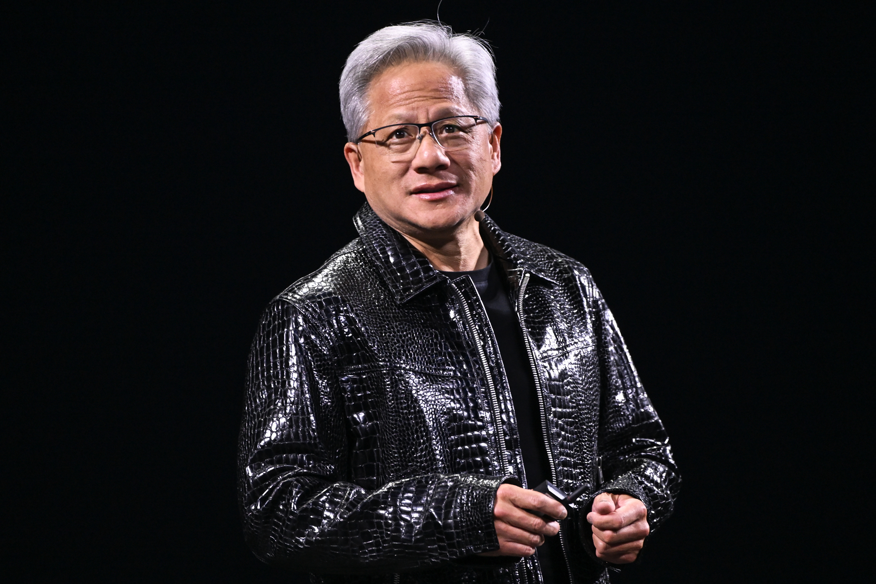 A person is wearing a shiny, black, textured jacket and glasses, standing against a dark background, holding a small object in their hands.