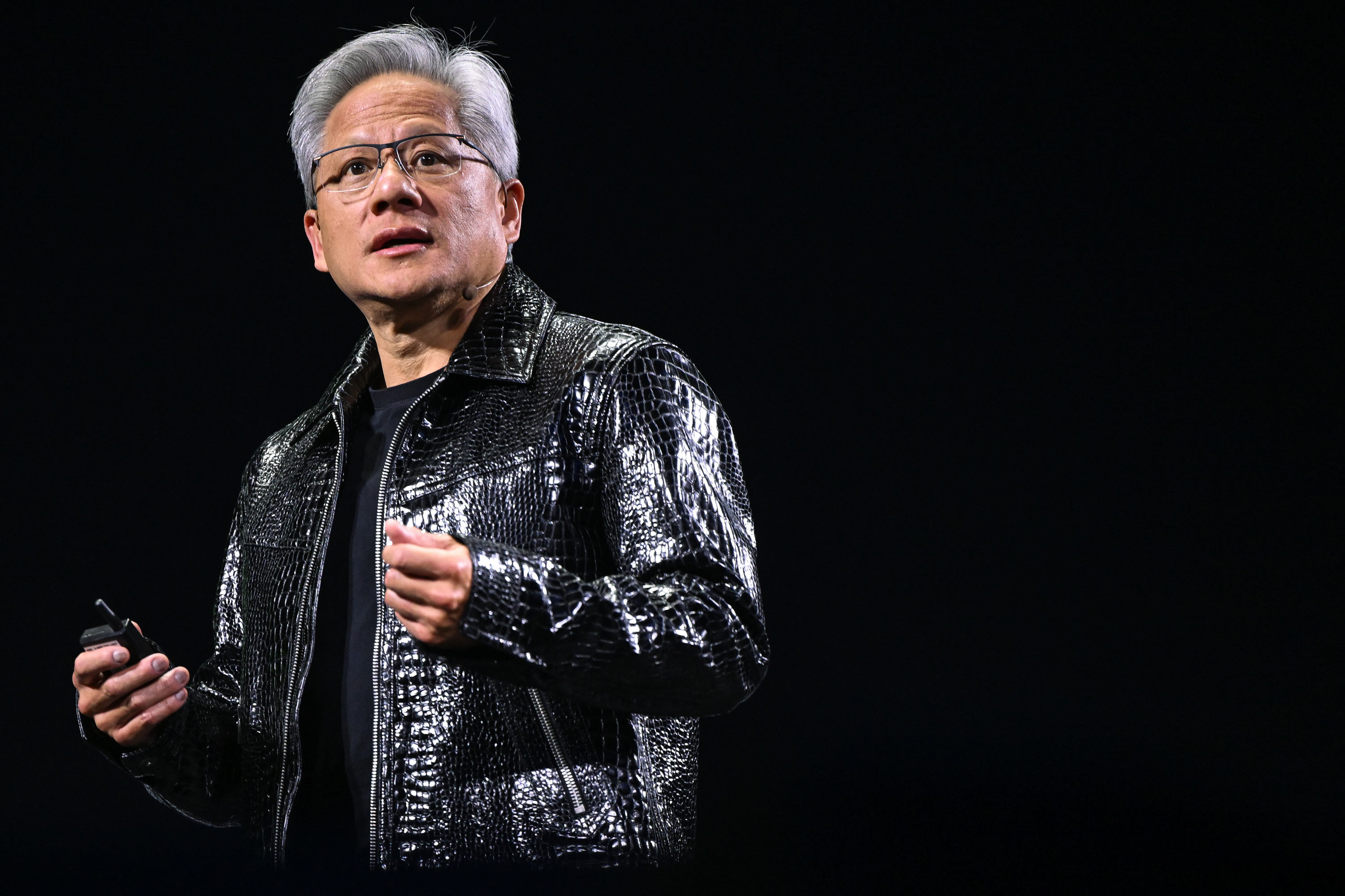 A person with short gray hair and glasses is speaking on stage, wearing a shiny black jacket and holding a small device, with a dark background.