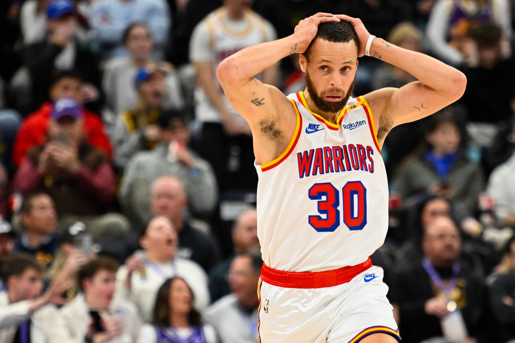 A basketball player wearing a Warriors jersey with the number 30 looks surprised, with hands on his head. The audience in the background seems engaged.