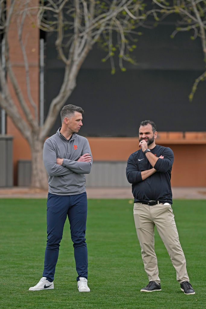 Two men stand on grass, each with arms crossed. The one on the left wears a gray top and blue pants, while the one on the right has a black shirt and khaki pants.