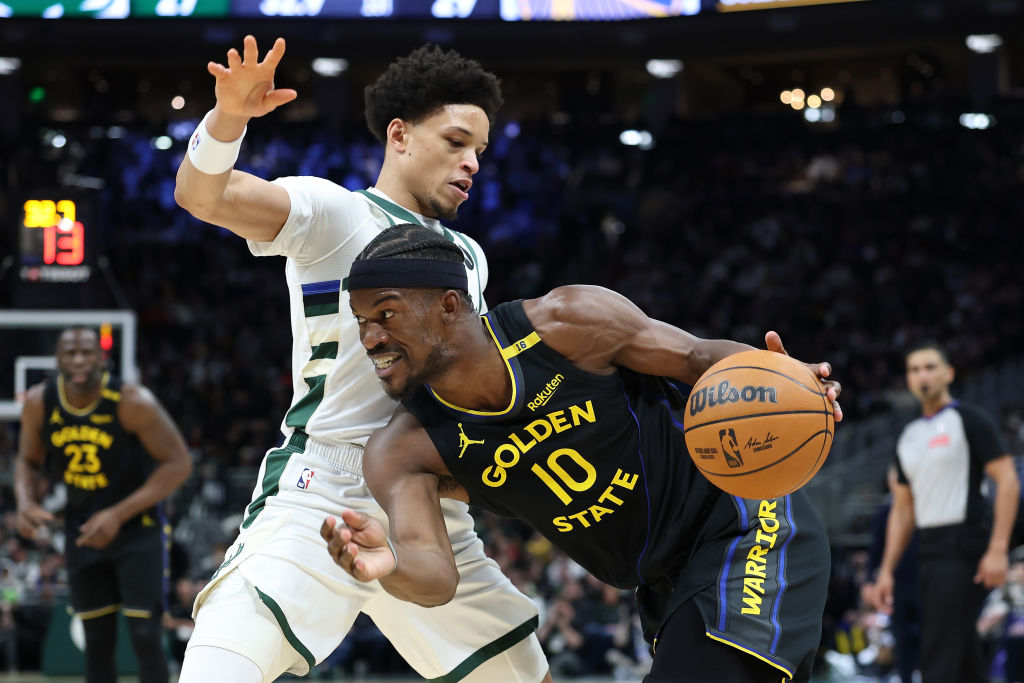 Two basketball players are in action. One in a Golden State jersey is dribbling the ball while the other, in a white jersey, defends closely. An official is in the background.