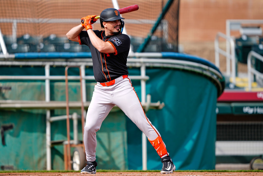 A baseball player stands ready to bat, wearing a black and orange uniform with a helmet and gloves. The background shows batting cages and equipment.