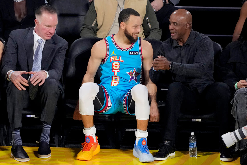 A basketball player in an &quot;All-Star&quot; jersey sits smiling between two men in suits. One man gestures while the other looks on. A water bottle is on the floor.