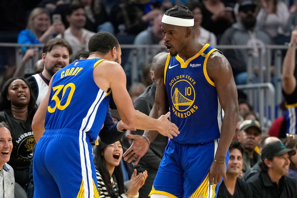 Two basketball players in blue Golden State Warriors uniforms are giving each other a handshake on the court, surrounded by cheering fans.