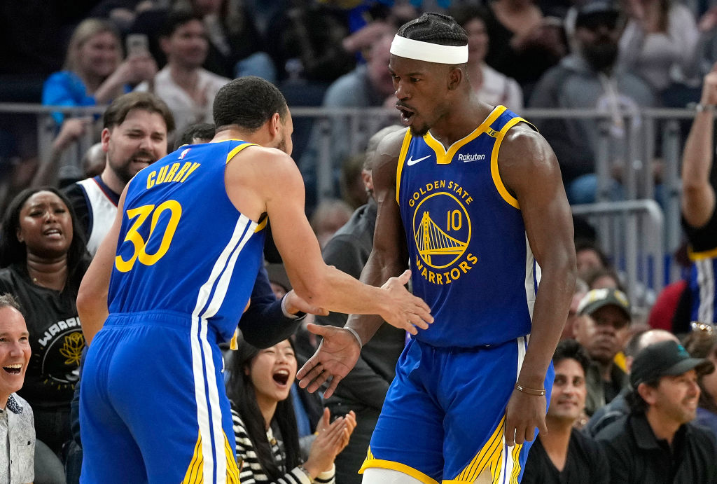 Two basketball players in blue Golden State Warriors uniforms are giving each other a handshake on the court, surrounded by cheering fans.