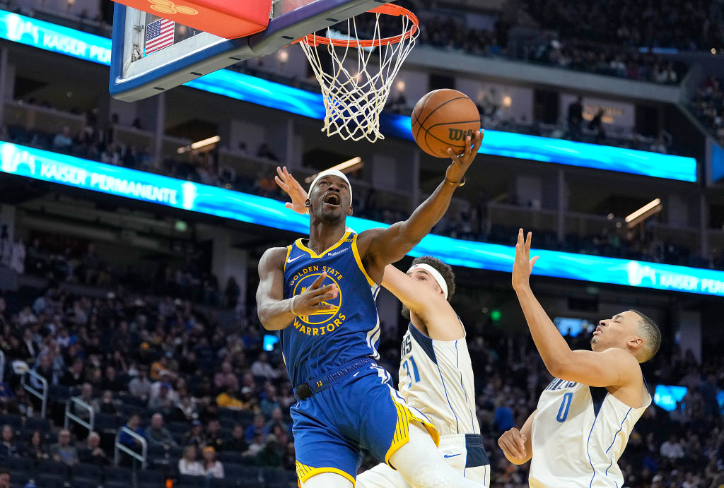 A basketball player in a Golden State Warriors jersey is driving to the hoop, with two defenders attempting to block him, in a busy, brightly lit arena.