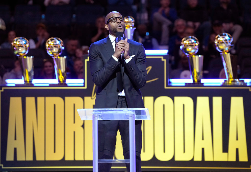 A person in a suit speaks at a podium with five trophies behind them, against a backdrop with the name &quot;Andre Iguodala&quot; prominently displayed.