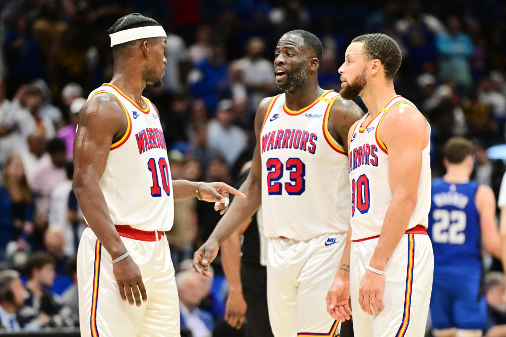 Three basketball players in white &quot;Warriors&quot; jerseys are talking on the court. One is wearing a headband, and number 23 is speaking in the middle.