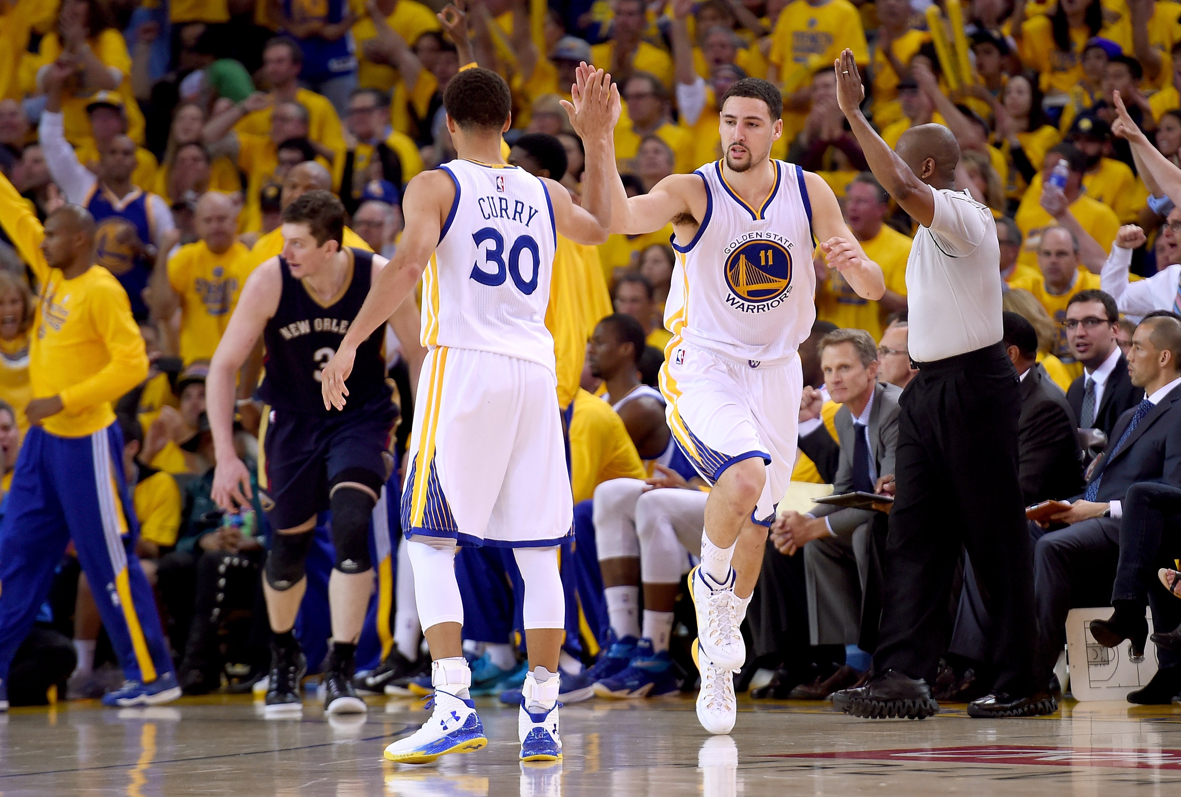 Two basketball players in white jerseys high-five on the court, while a referee signals nearby. A crowd wearing yellow cheers in the background.