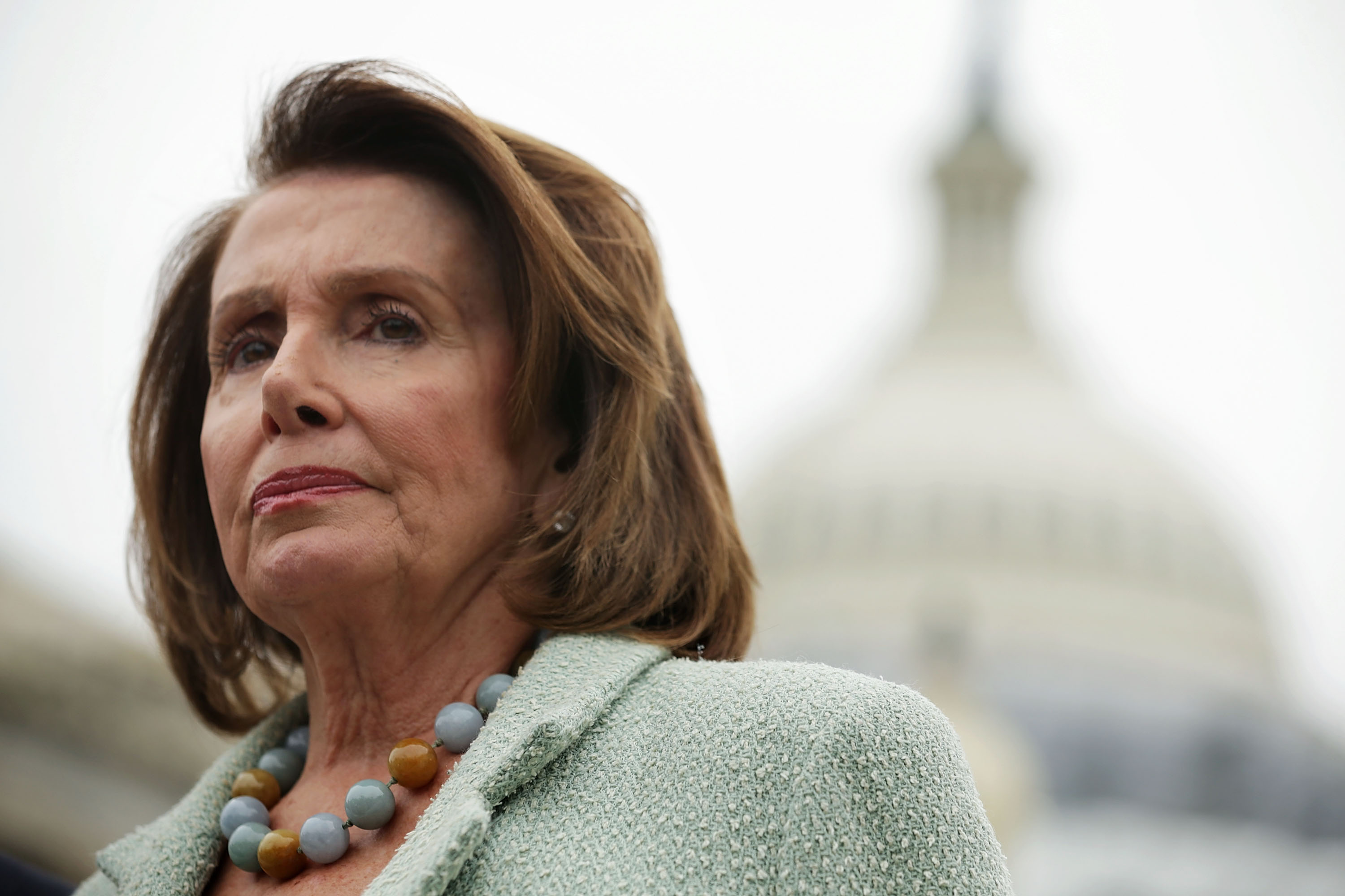 A woman with short brown hair, wearing a beaded necklace and a textured jacket, stands in front of a blurred dome structure. Her expression is serious.