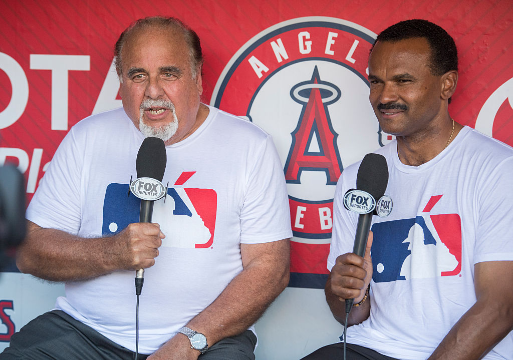 Two men are seated, each holding a Fox Deportes microphone. They wear matching white shirts with an MLB logo. A red Angels baseball background is behind them.