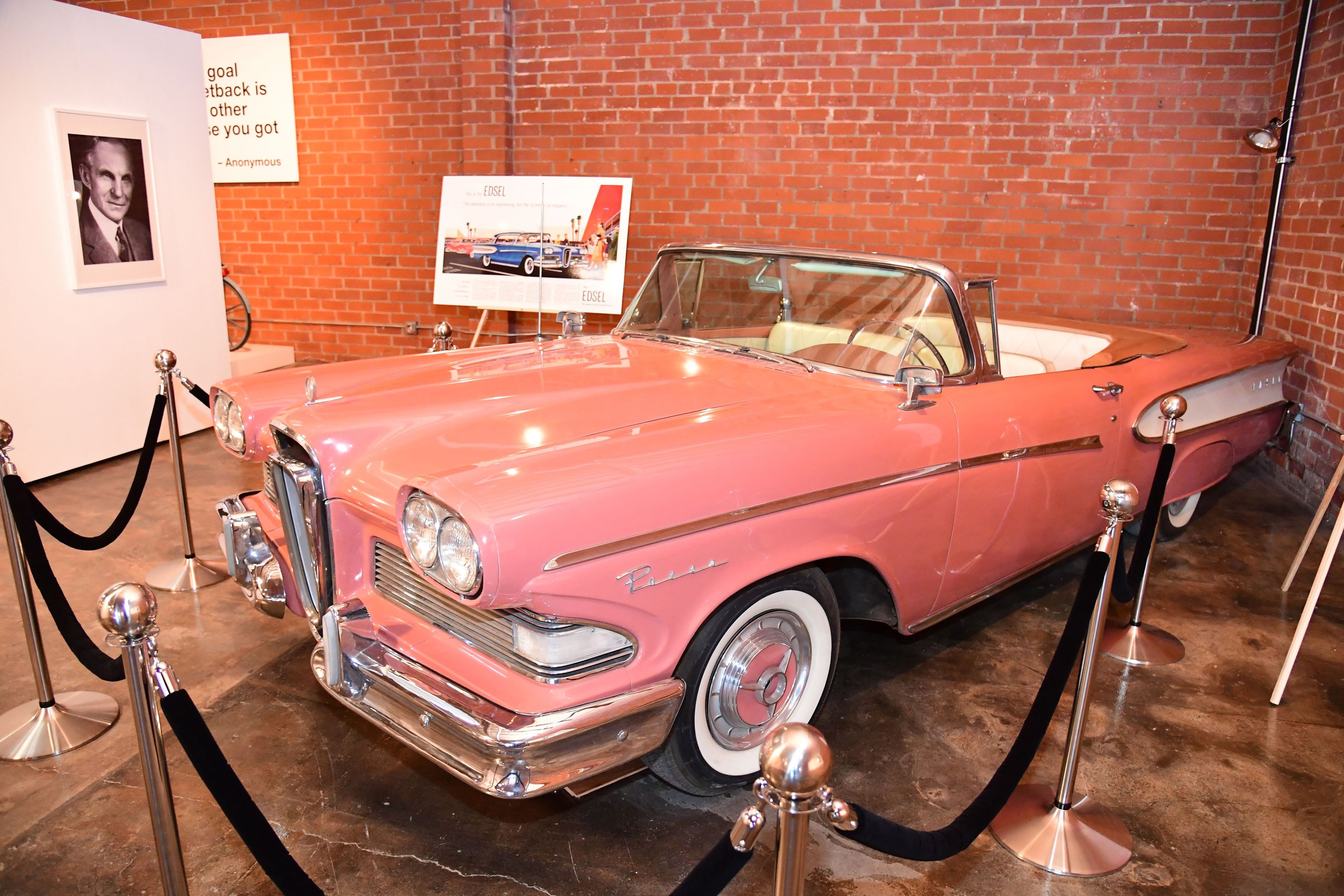 A pink vintage convertible car is displayed in a museum, roped off with black stanchions. It sits near a brick wall with an old car advertisement.