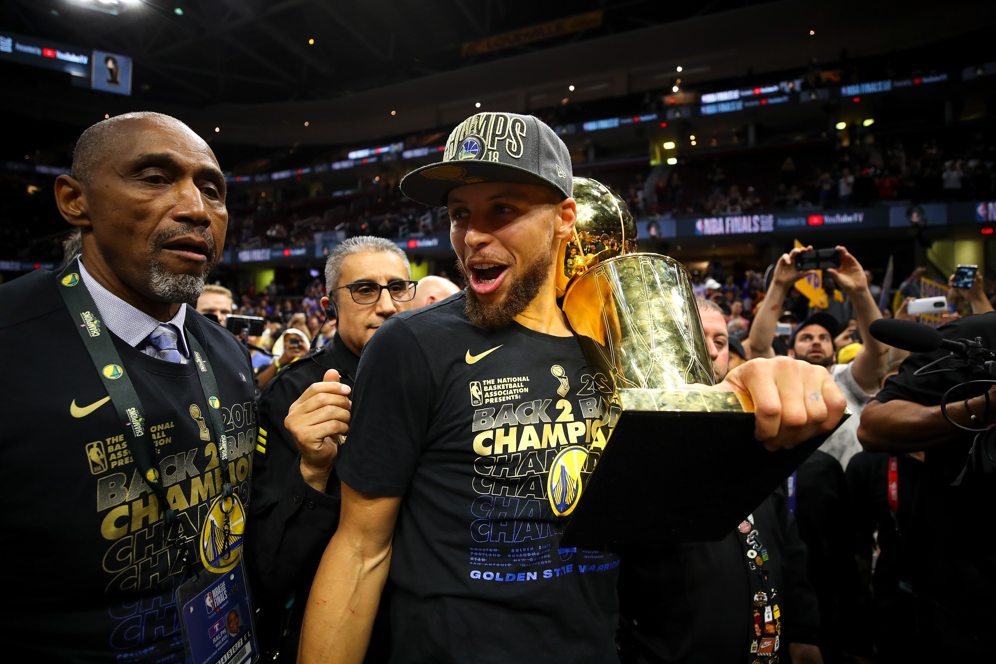 A basketball player joyfully holds a trophy, wearing a &quot;Back 2 Back Champions&quot; shirt and cap, surrounded by others in a lively sports arena.