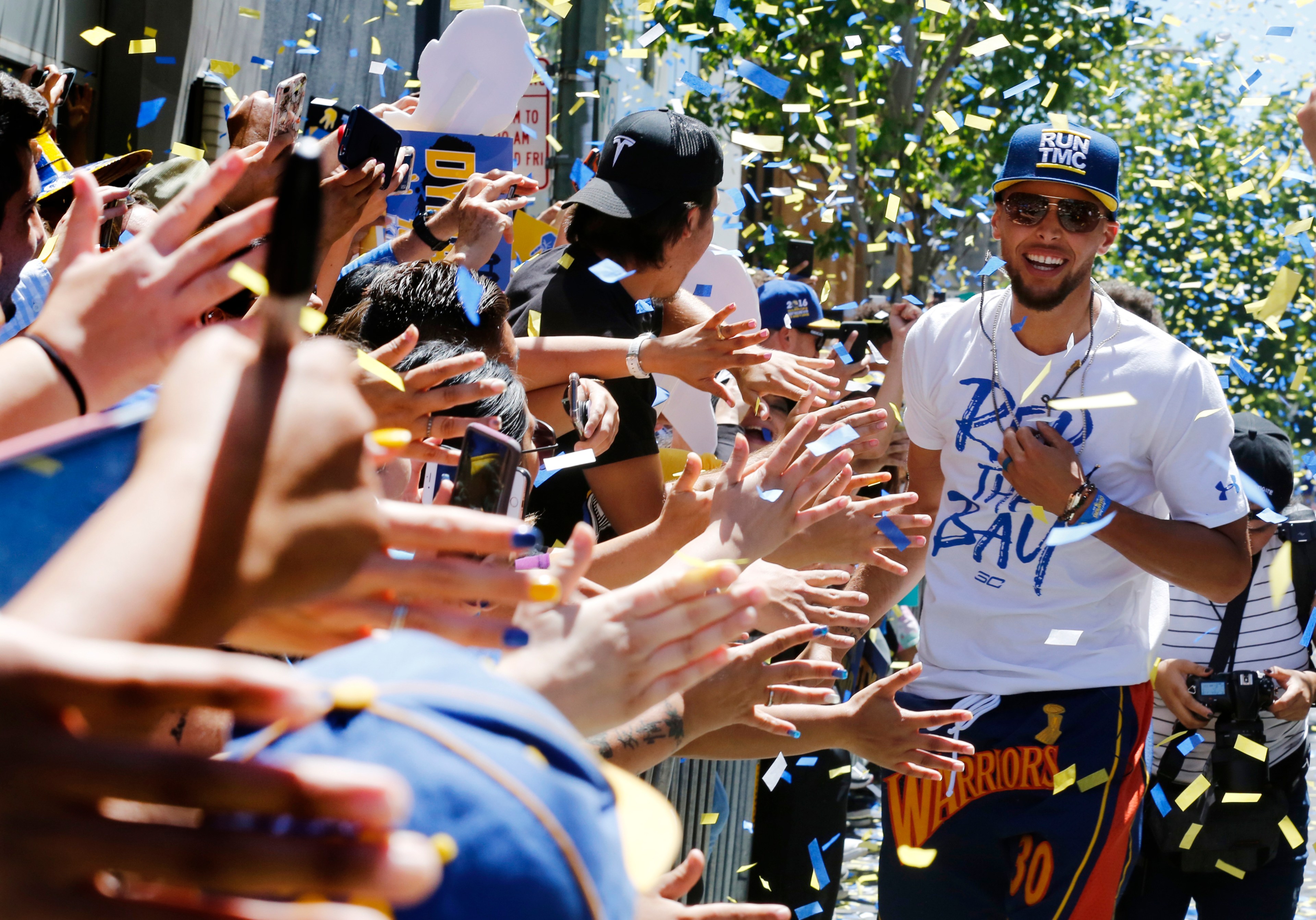 A man in a white shirt and cap smiles, walking through a crowd with arms outstretched, surrounded by cheering fans and falling confetti.