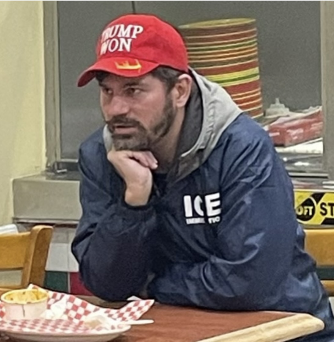 A man sits at a table wearing a red &quot;TRUMP WON&quot; hat and a blue jacket with &quot;ICE&quot; on it. He rests his chin on his hand. There's food in the foreground.