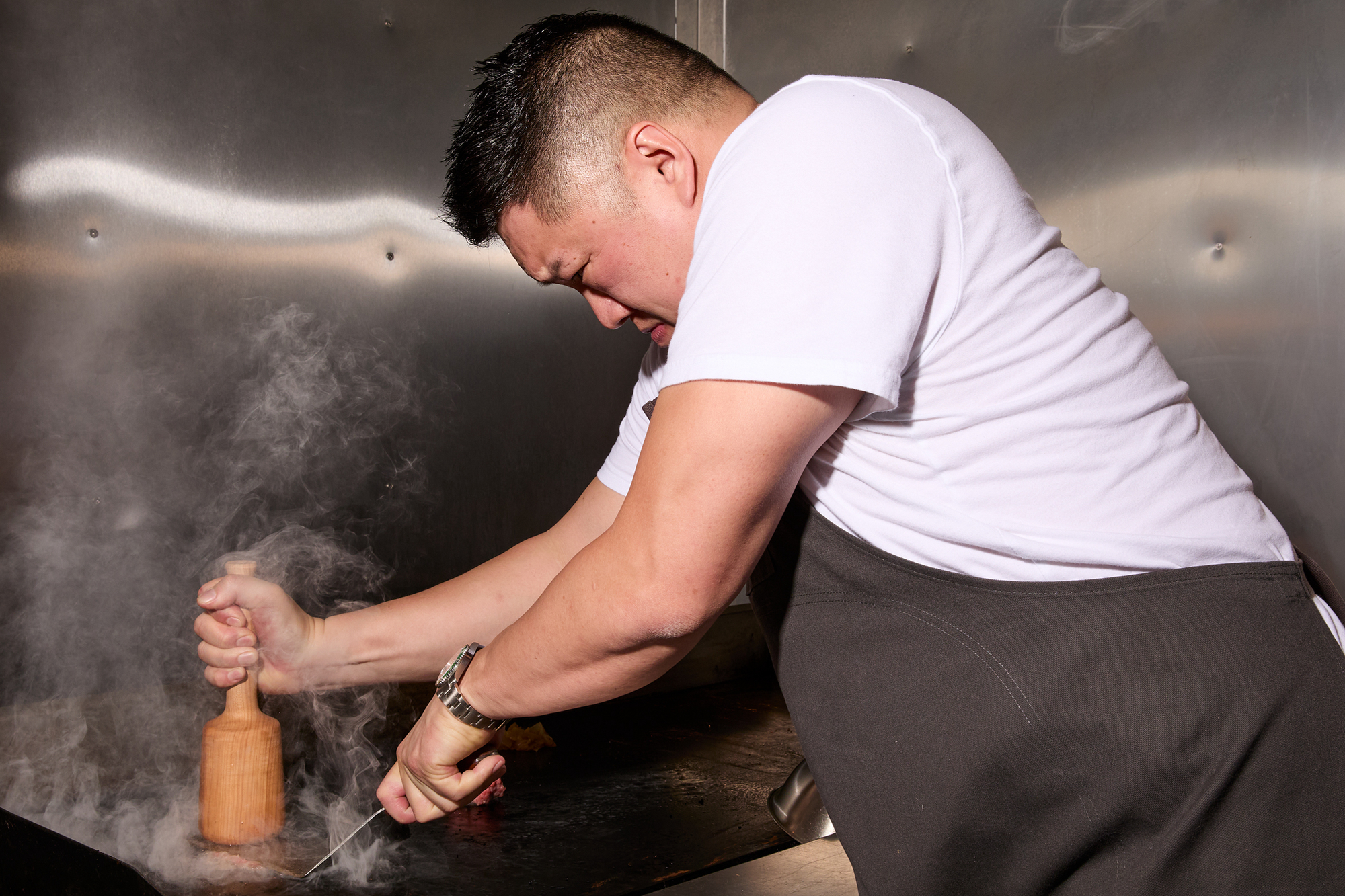 A person in a white shirt and dark apron is pressing a wooden tool onto a steaming hot griddle, concentrating intensely in a kitchen setting.