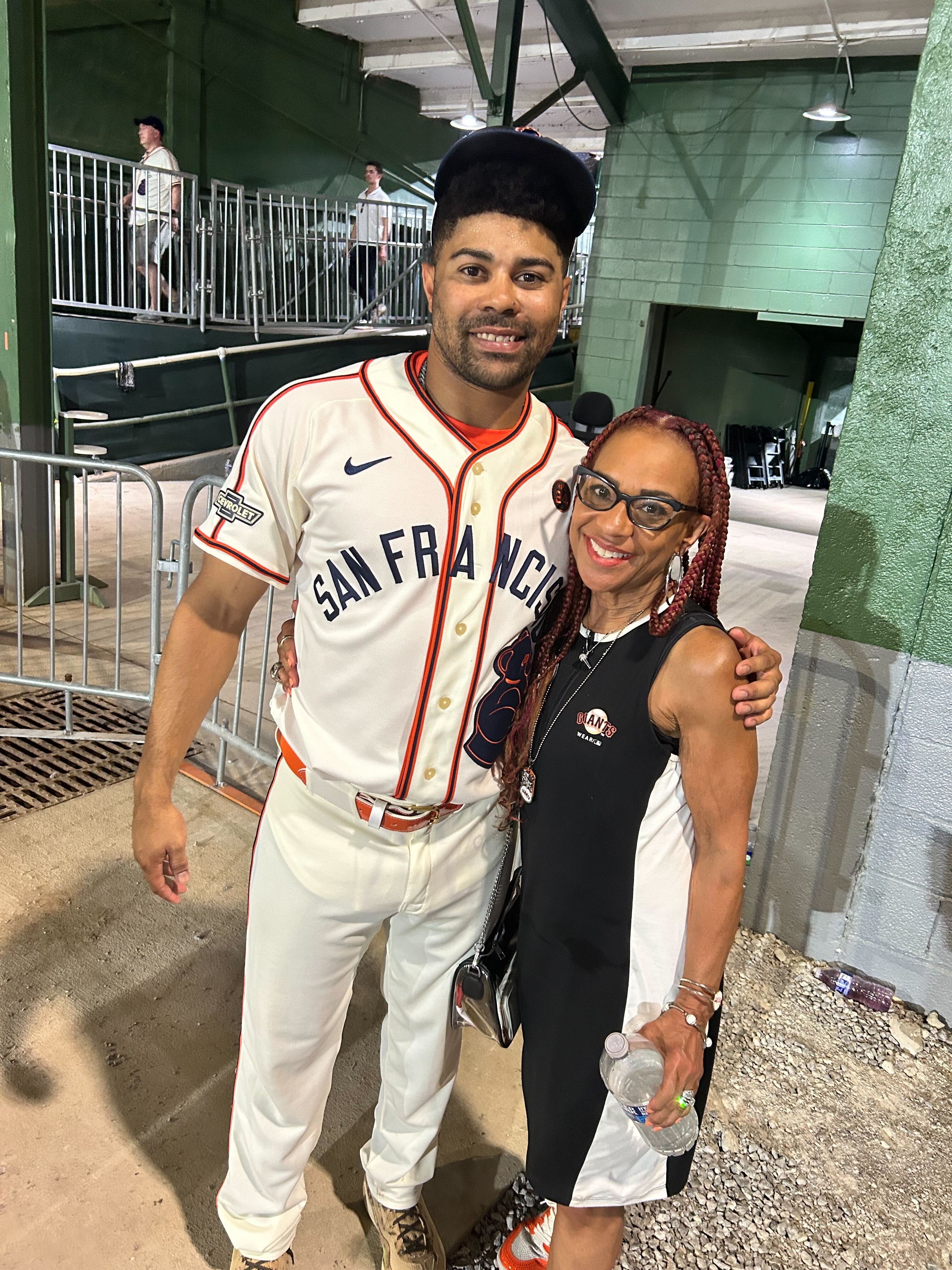 A baseball player in a &quot;San Francisco&quot; uniform poses with a smiling woman in a black and white dress inside a stadium area. The ground is gravelly.