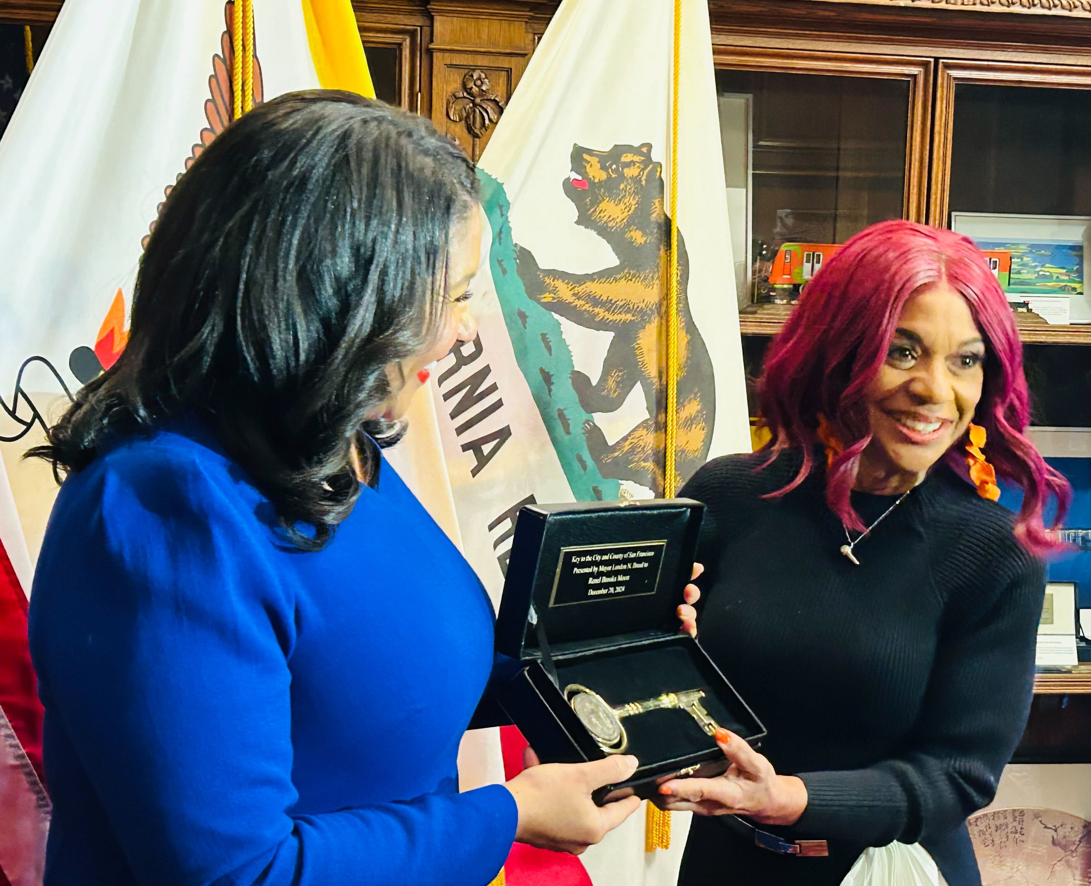 Two women are shown; one holds an open box containing a ceremonial key. Flags are visible in the background, and they both appear to be smiling.