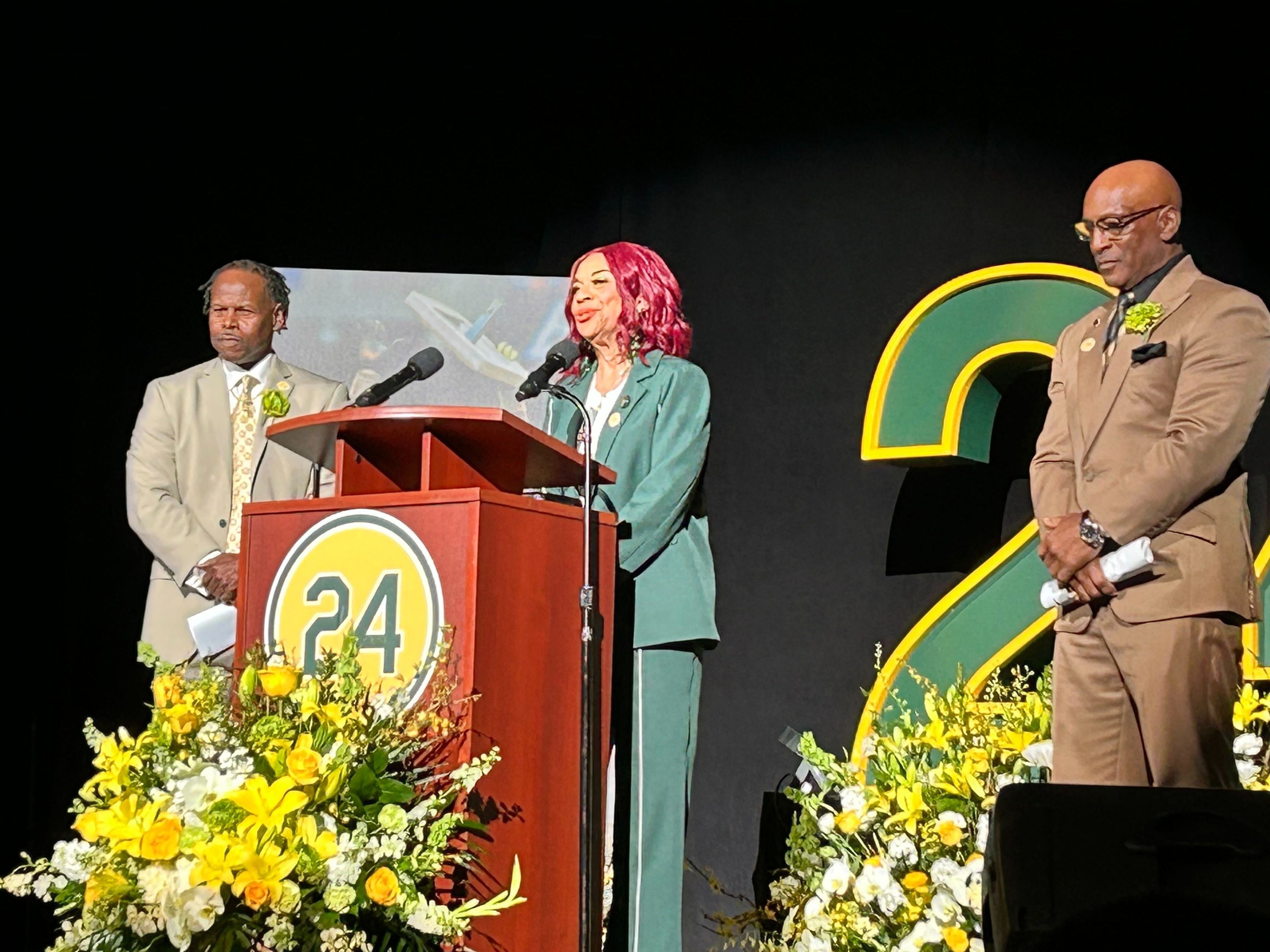 Three people stand on a stage, two men and a woman in green. They're near a podium marked &quot;24,&quot; surrounded by yellow and white floral arrangements.