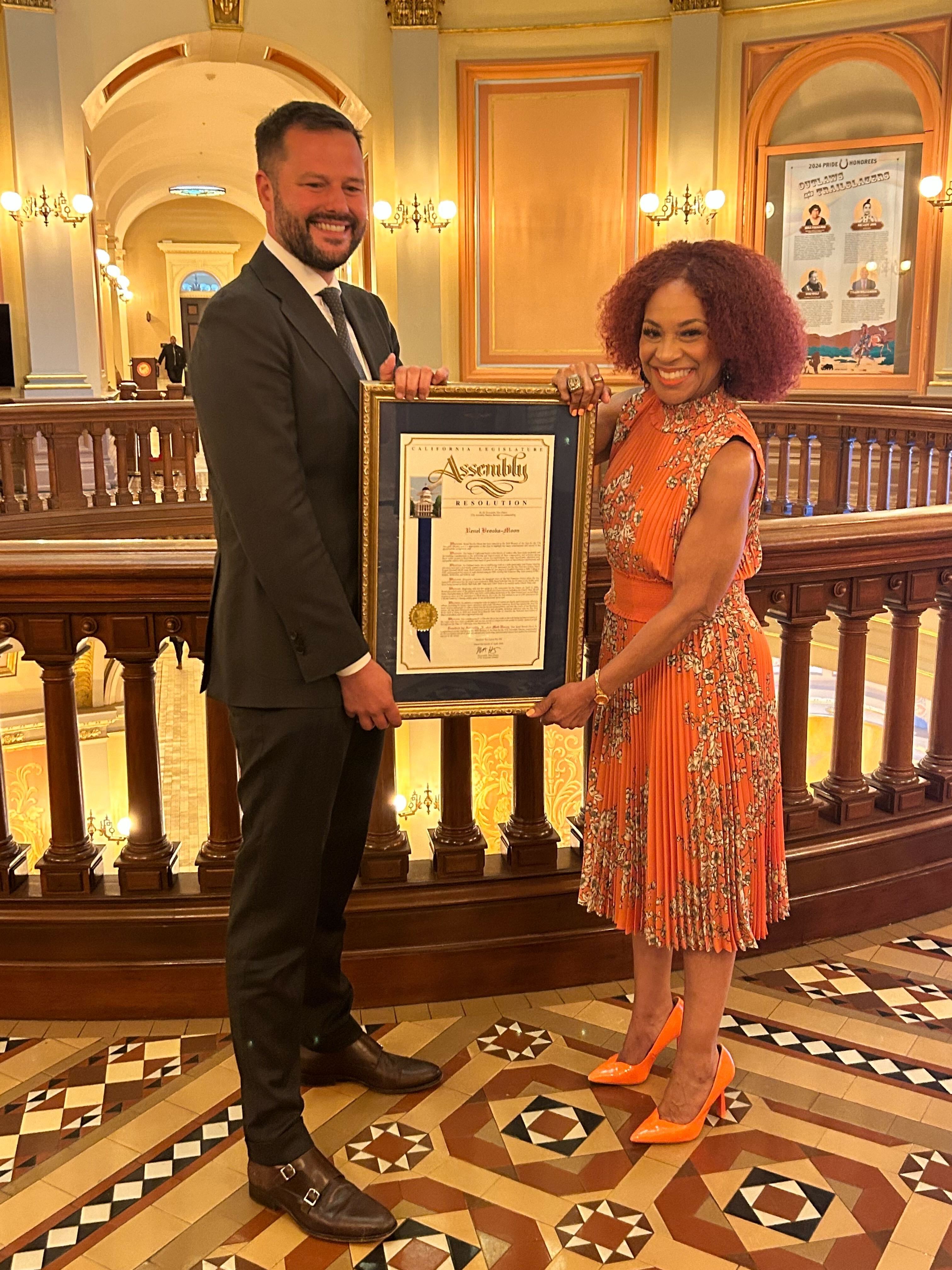 Two people are standing, smiling, and holding a framed document. They're in a decorative interior with an ornate tiled floor, arches, and lit chandeliers.